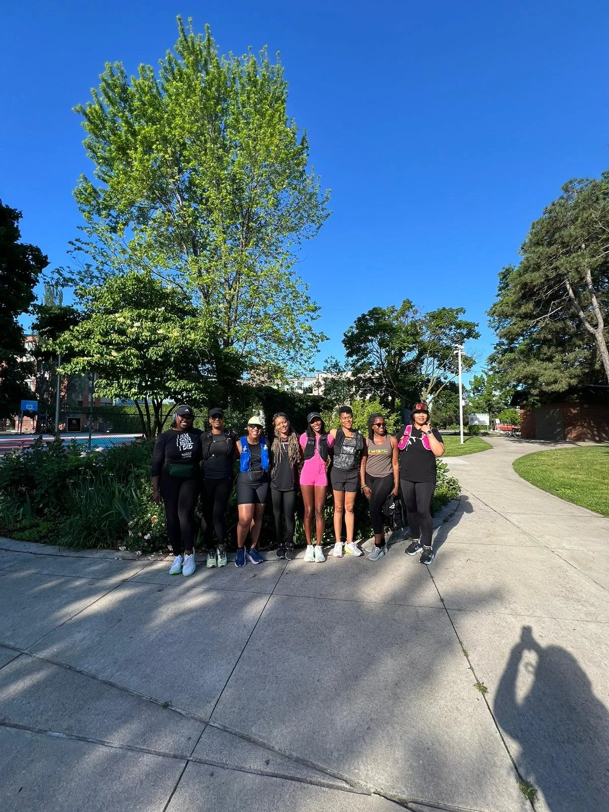 A group of eight women wearing athletic clothing and backpacks standing on a sidewalk in a park with green trees and a blue sky.
