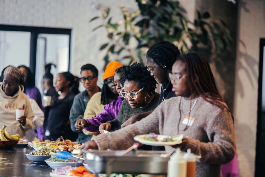 Group of diverse women standing in line at a buffet table, serving themselves food during a gathering in an indoor setting.