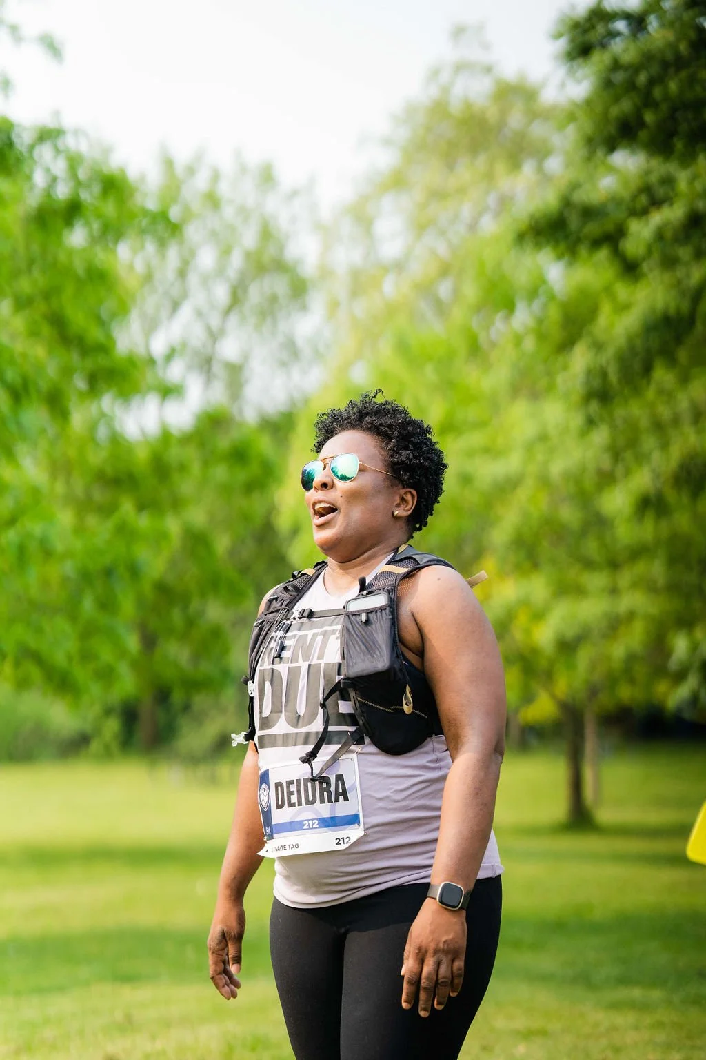 Woman with sunglasses, wearing a race bib, and a running vest, smiling outdoors with greenery in the background.