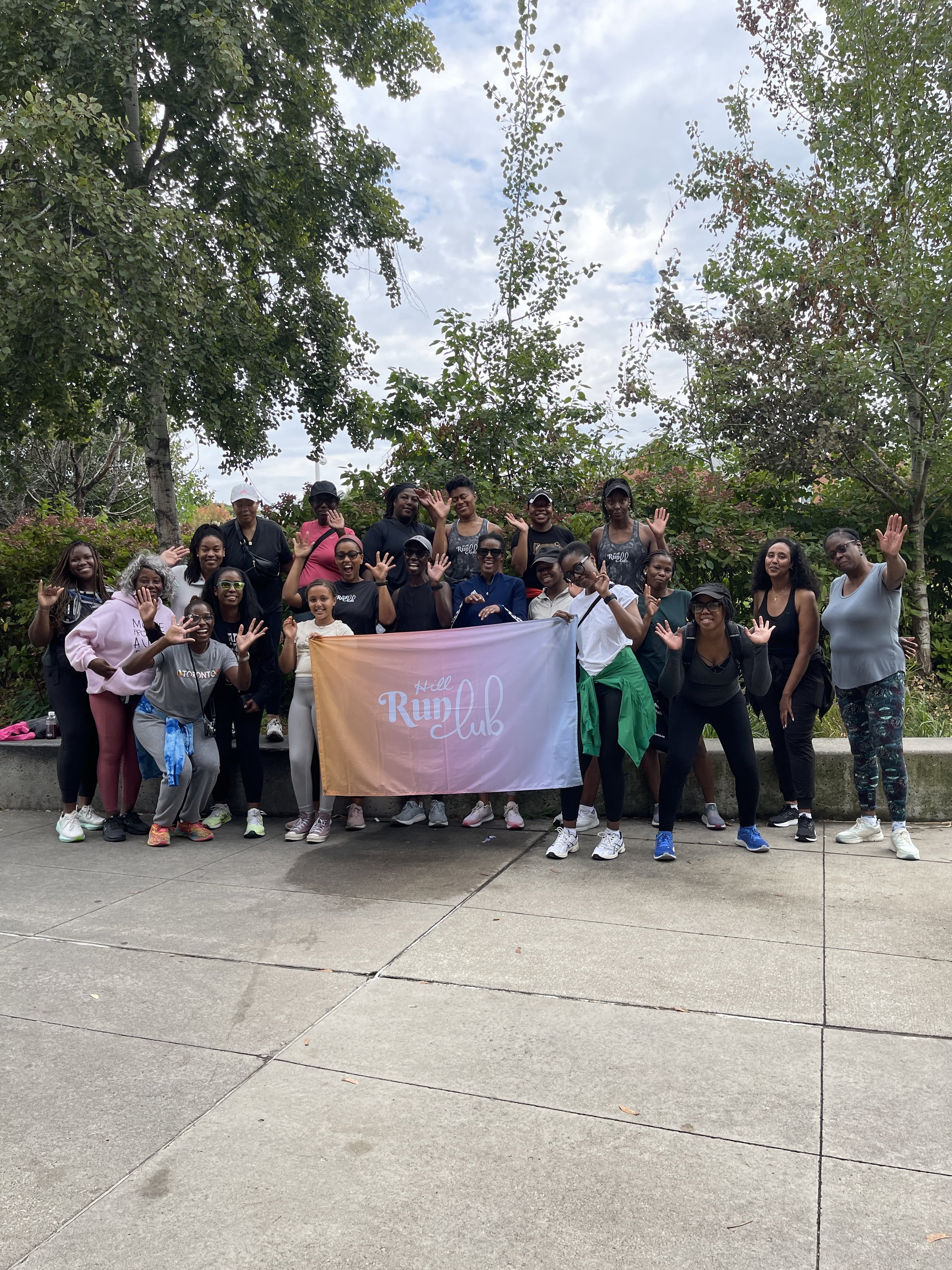A group of women holding a banner that reads 'Hill Run Club,' standing outdoors on a sidewalk with trees and cloudy sky in the background, waving and smiling at the camera.