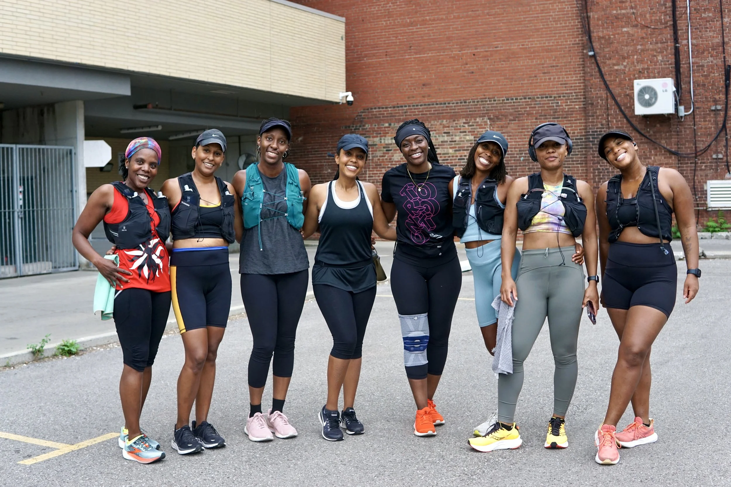A group of eight women dressed in athletic wear, standing outdoors in front of a red brick building, smiling and posing for a photo after a workout.