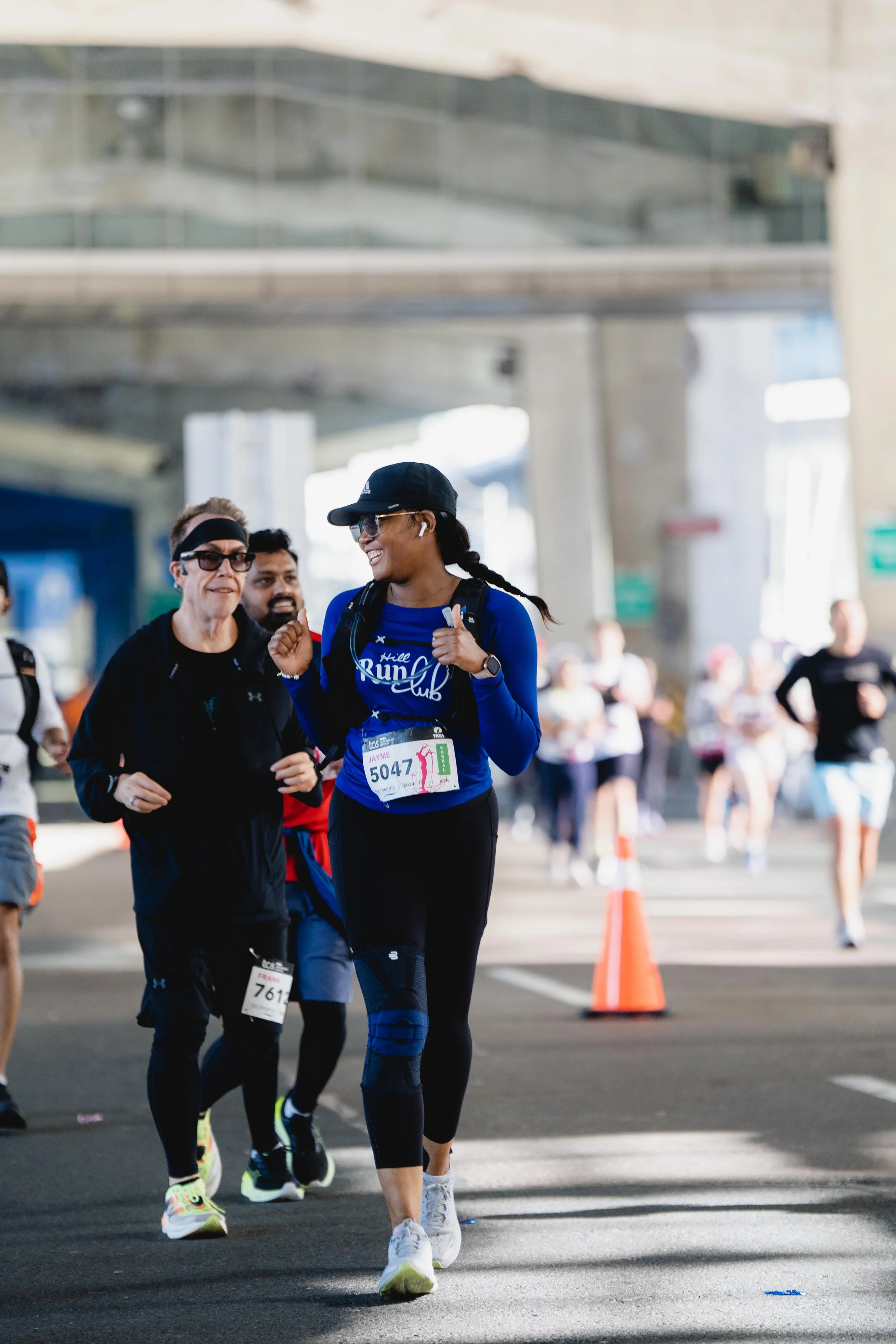 A group of runners participating in a marathon, with a woman in a blue running outfit, race bib number 5047, smiling and giving a thumbs-up, under a bridge with other runners and spectators in the background.
