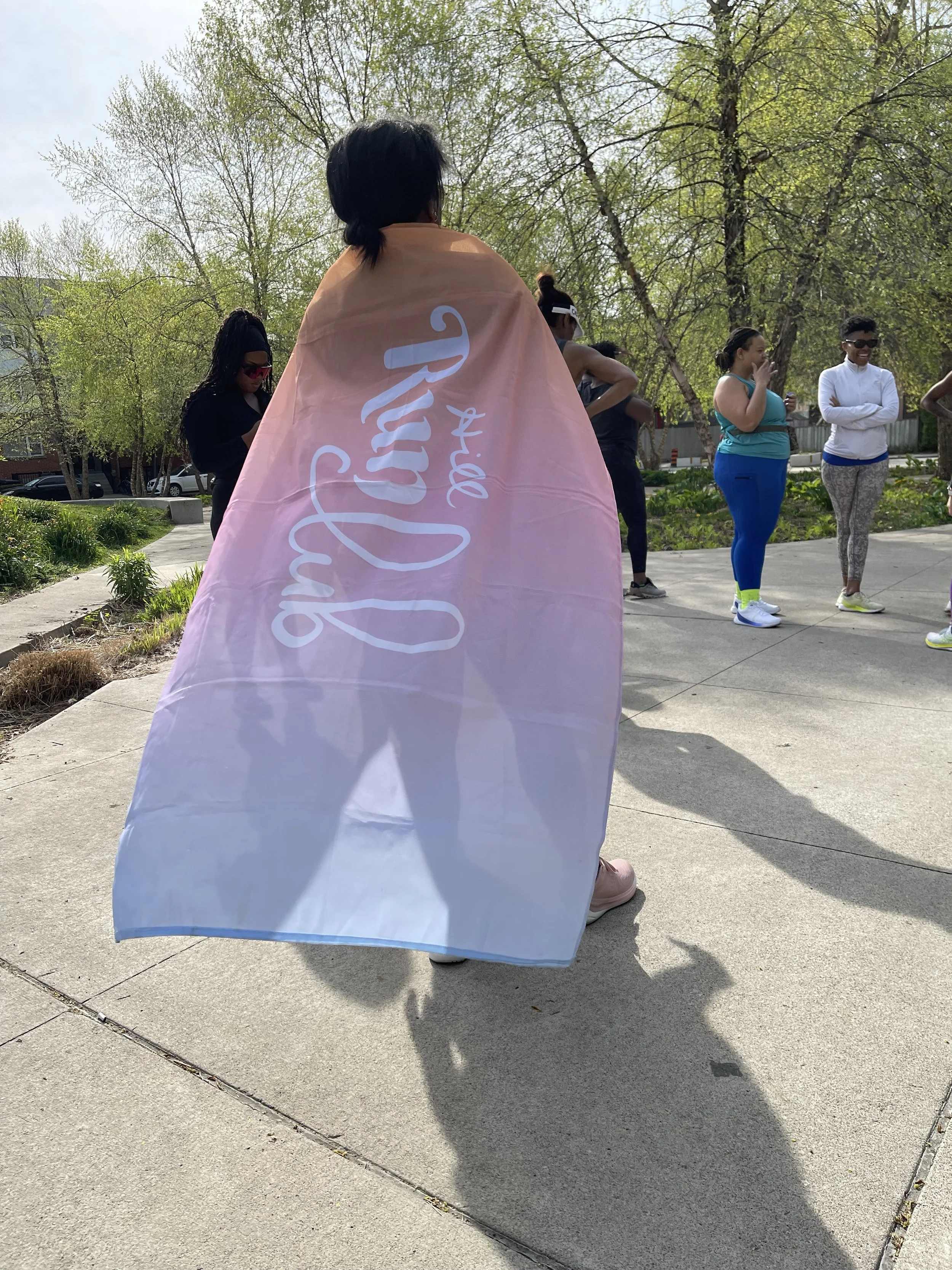 A group of people standing outdoors on a sidewalk, with one person in the foreground draped in a pink and blue gradient cape that says "Sorry Not Sorry" in cursive.