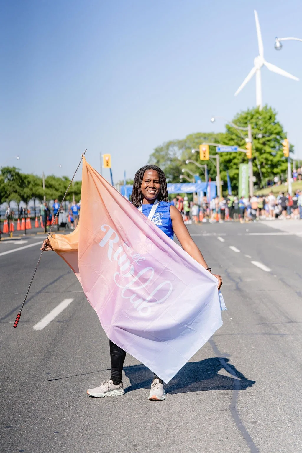 A woman holding a pink and white flag with 'The Royal Club' written on it, standing in the middle of a street during an outdoor event with a crowd and wind turbines in the background.