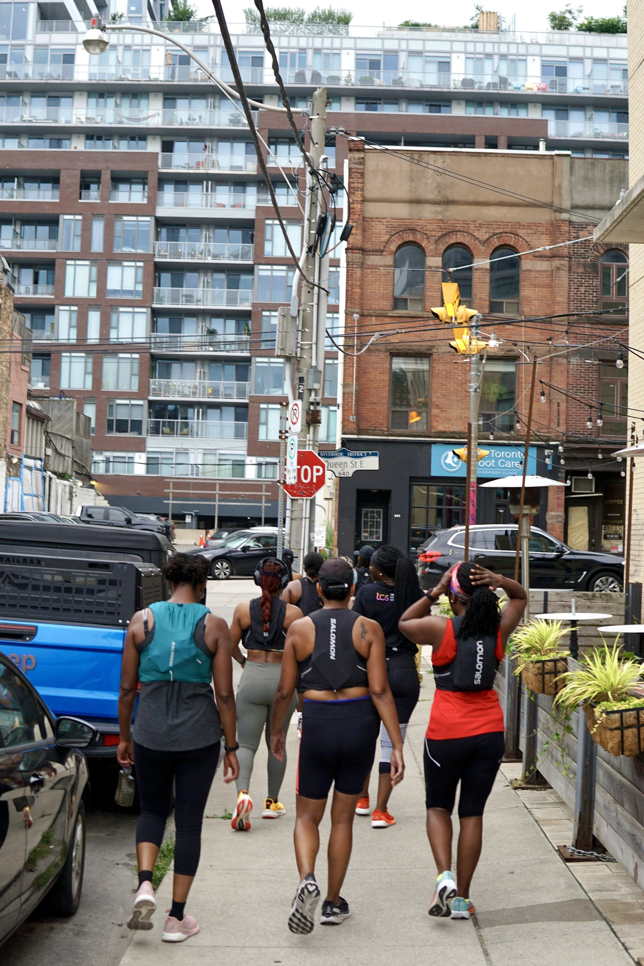 Group of women in athletic wear walking in an urban street, with residential and commercial buildings in the background, during daytime.