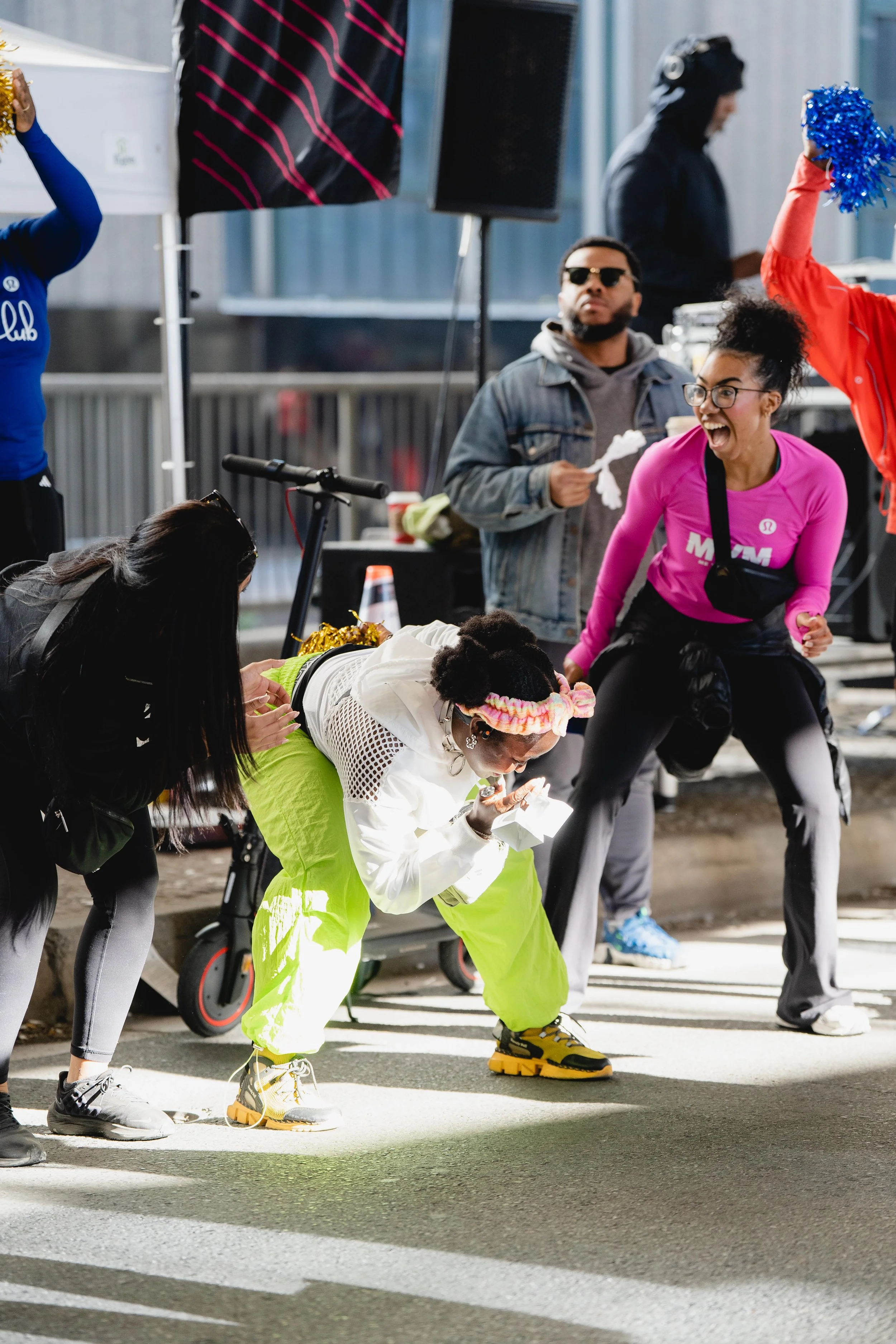 People celebrating at a marathon event, with one woman in neon yellow pants and a white jacket leaning forward, blowing a kiss, and others cheering around her.