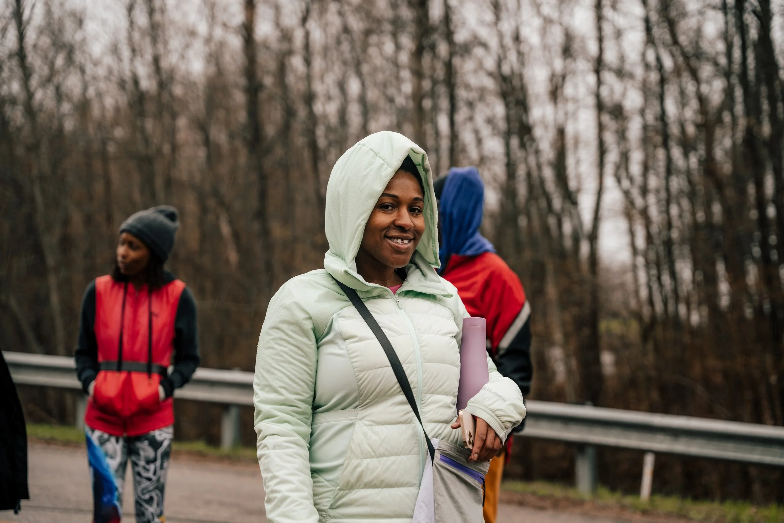 A group of people dressed in outdoor sportswear, including jackets and hats, standing on a roadside in a wooded area on a cloudy day.