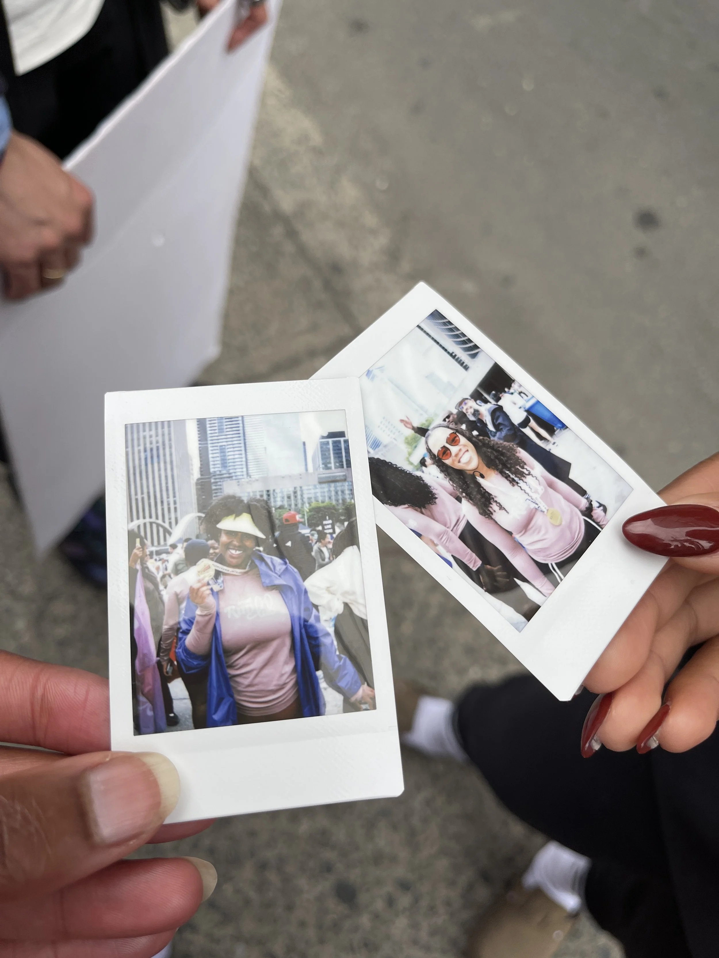 Two women holding up two polaroid photos with city buildings in the background, people in a crowd, and smiling faces.