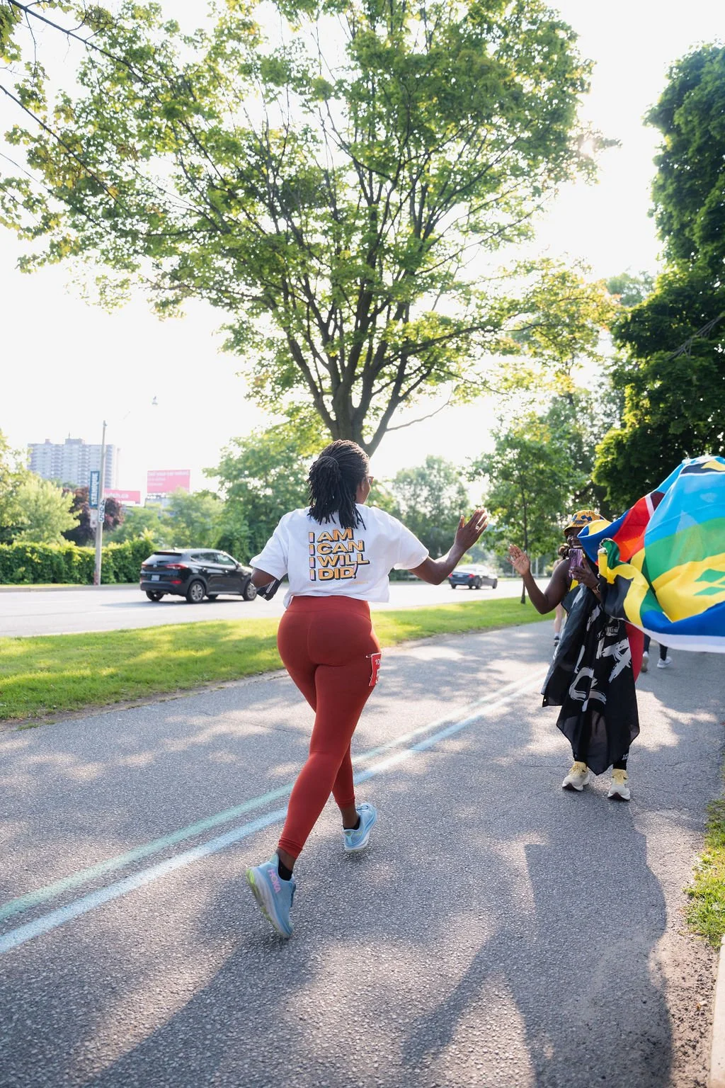 Woman running on a sidewalk during a race or event, high-fiving a supporter holding a rainbow flag, under a large tree with sunlight filtering through leaves.