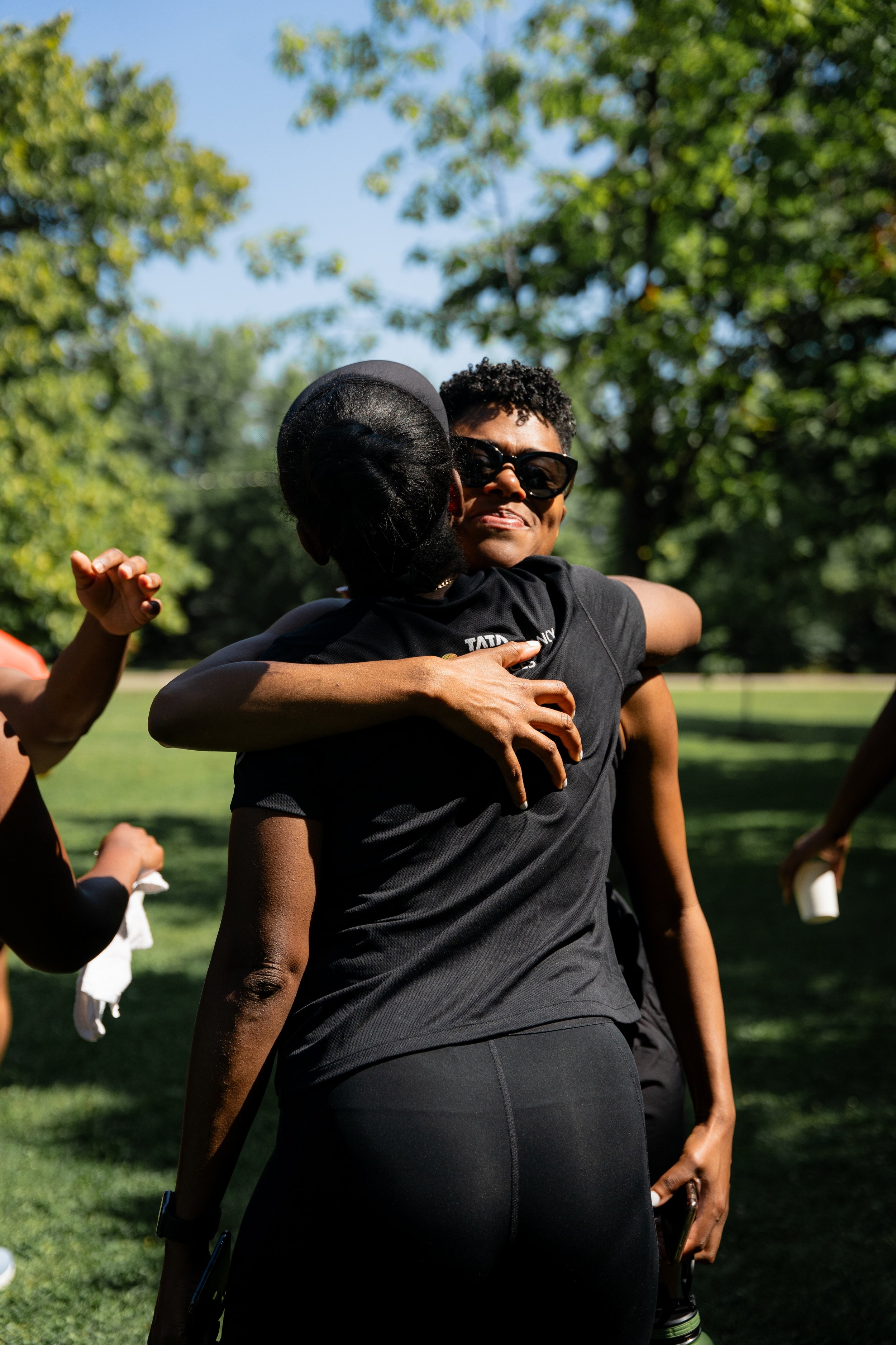 Two women hugging outdoors on a sunny day, with trees and grass in the background.