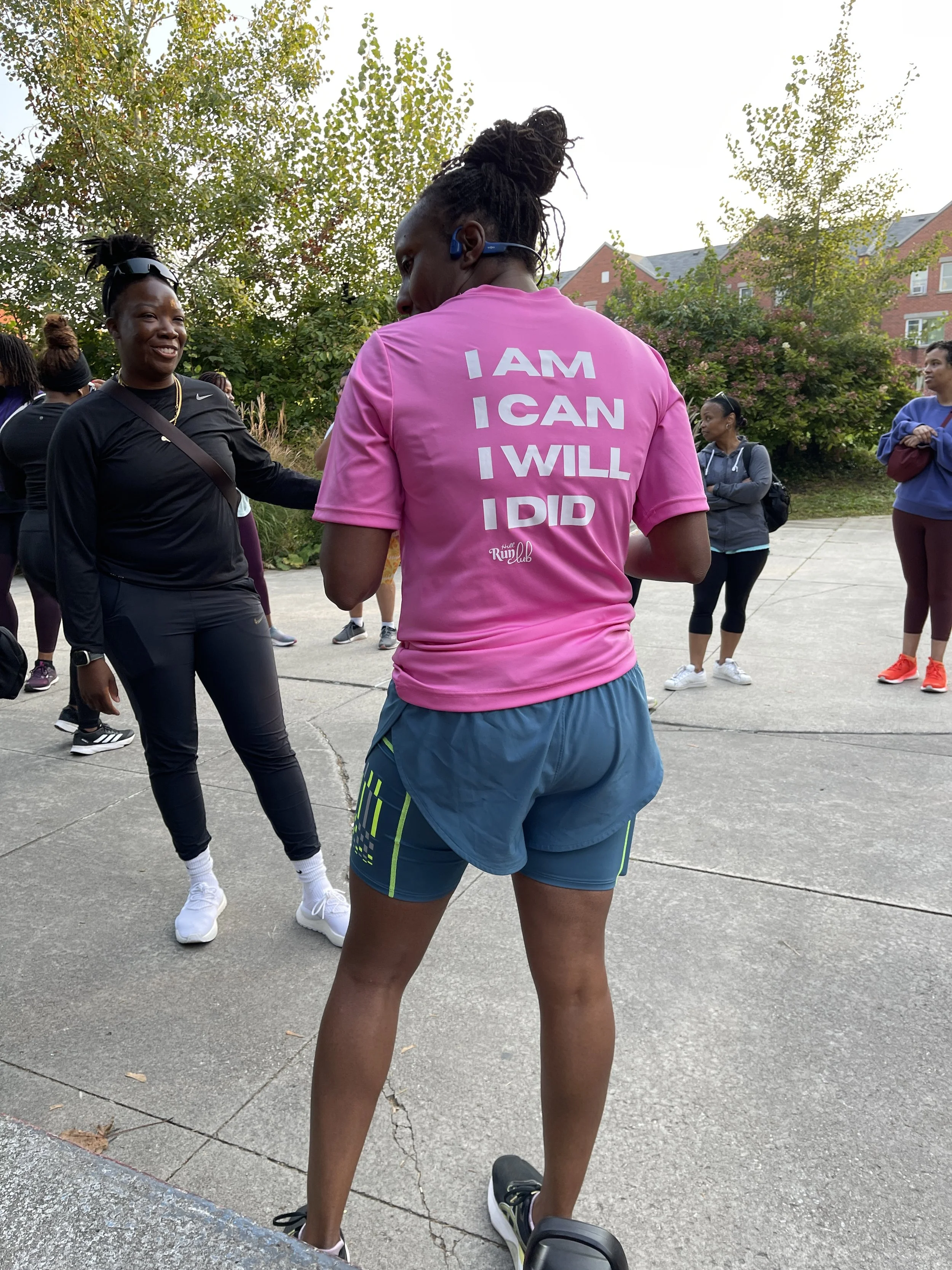 A group of women gathered outdoors, wearing athletic clothing, with trees and residential buildings in the background. One woman in the foreground has her back to the camera and is wearing a pink T-shirt with motivational words and blue shorts.