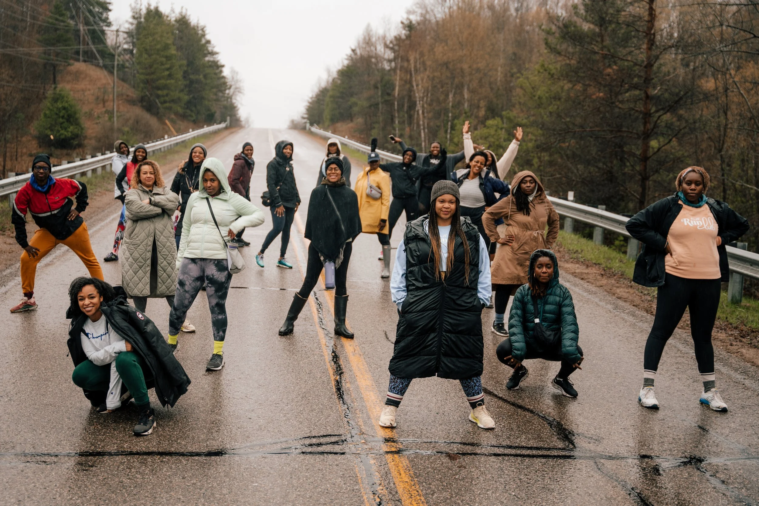 Group of women standing on a wet road with trees and hills in the background, wearing jackets and casual clothing.