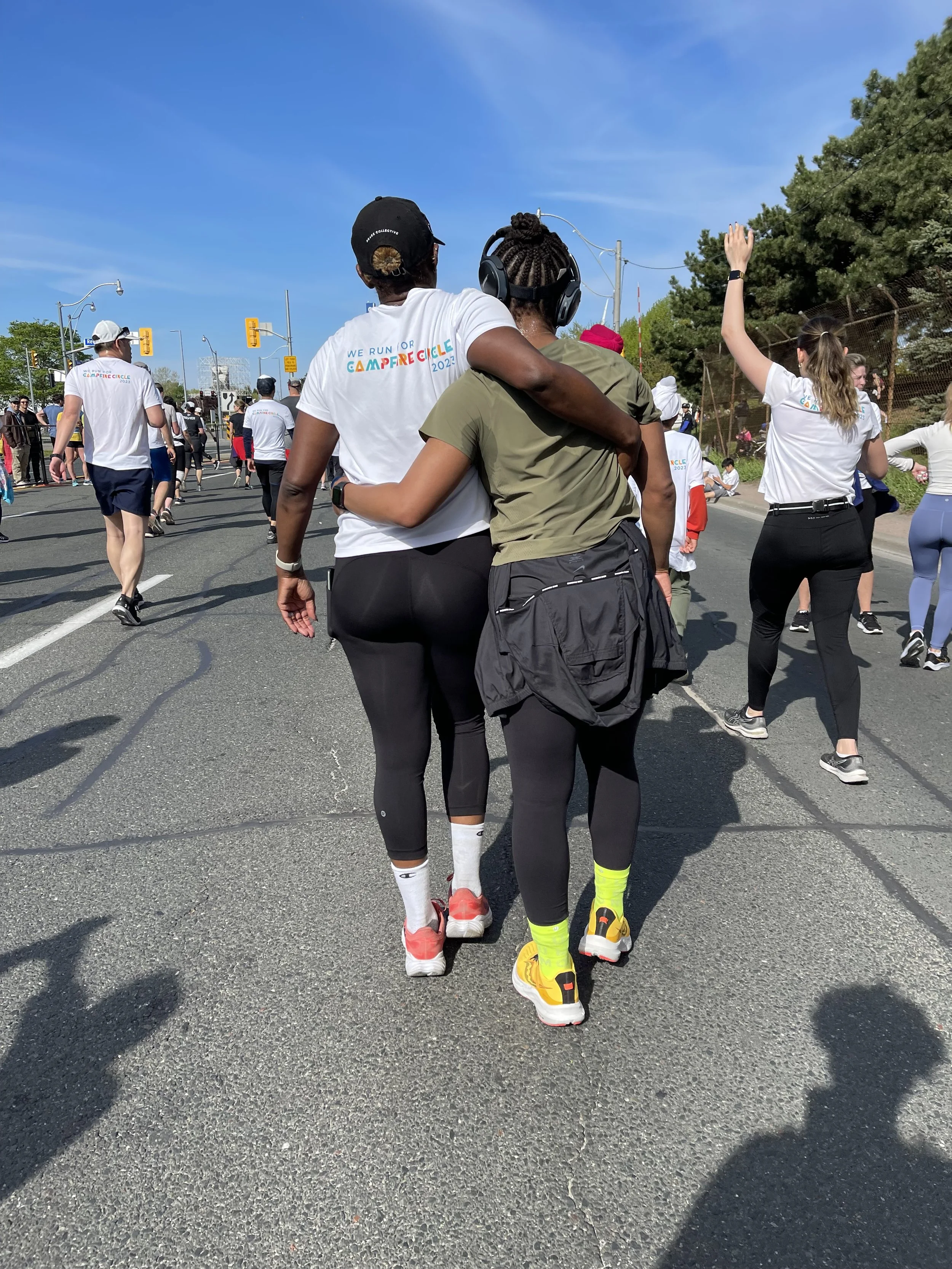 Two women walking arm in arm during a marathon event, surrounded by other runners and spectators on a sunny day.