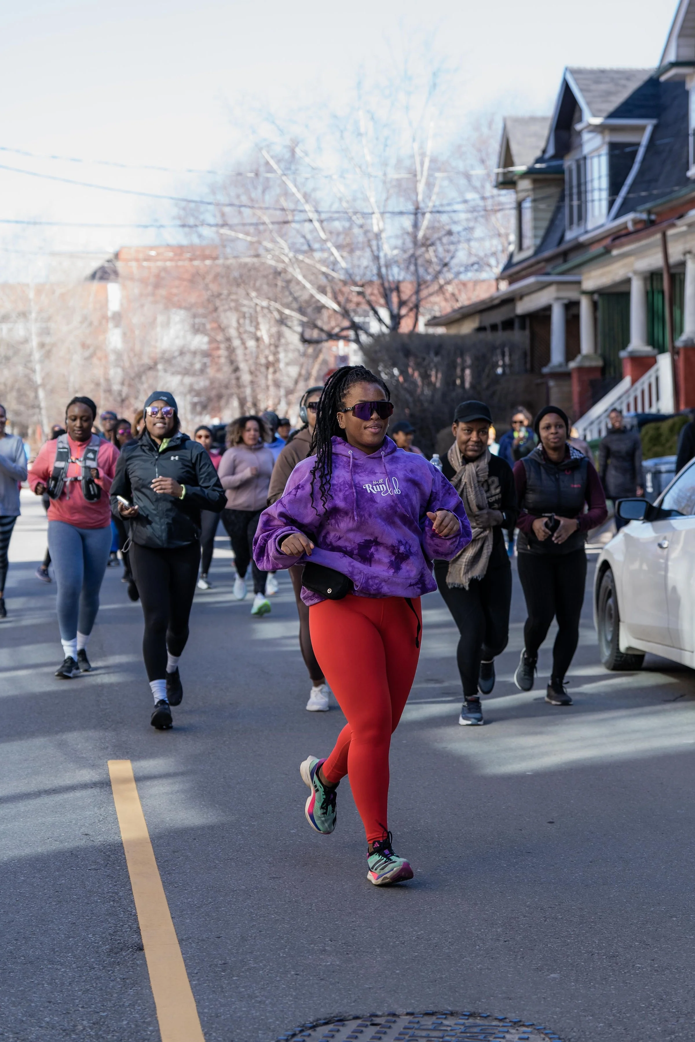A group of people participating in a community run or walk event on a residential street, with one woman in bright red leggings and purple hoodie leading the pack.