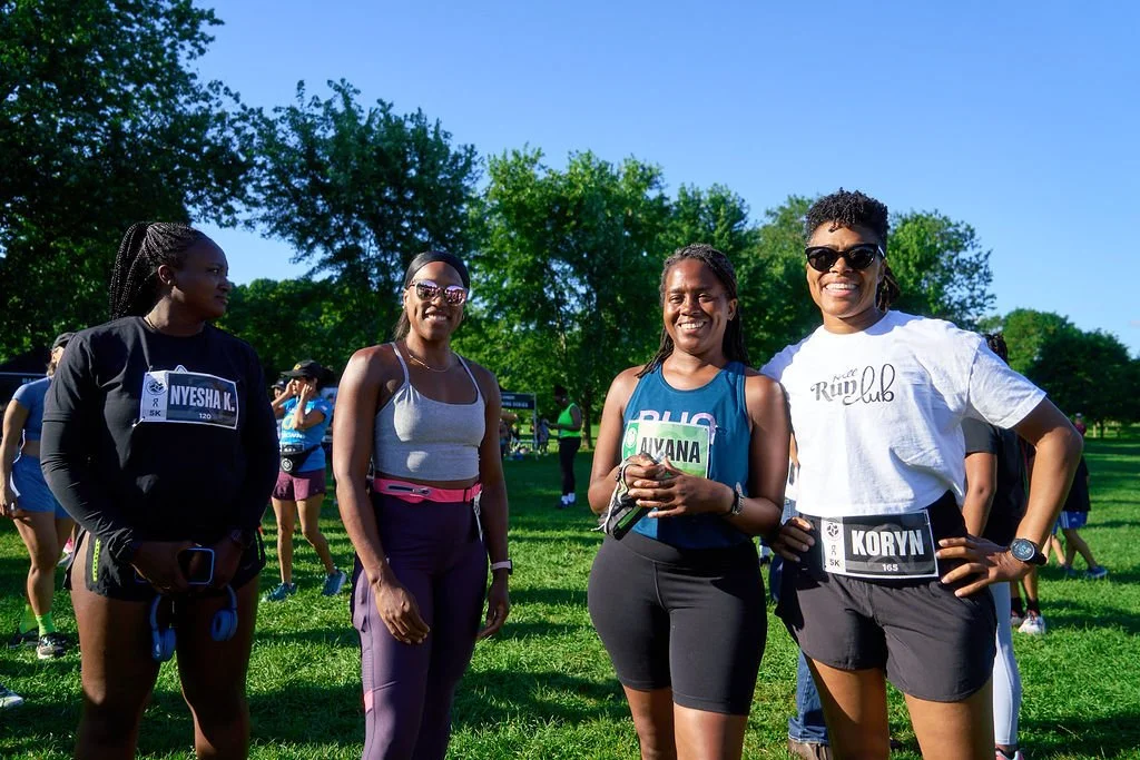 Four women standing together outdoors on a sunny day, participating in a running event, wearing athletic clothes and race bibs.