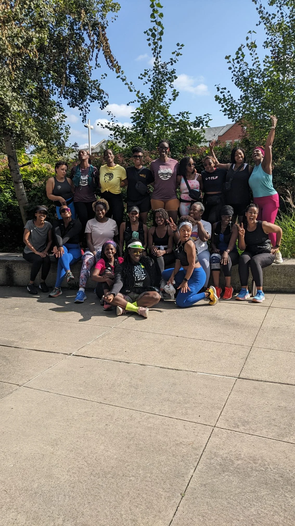 Group of women and girls outdoors on a sunny day, posing for a photo in front of trees and buildings, some making peace signs and smiling.