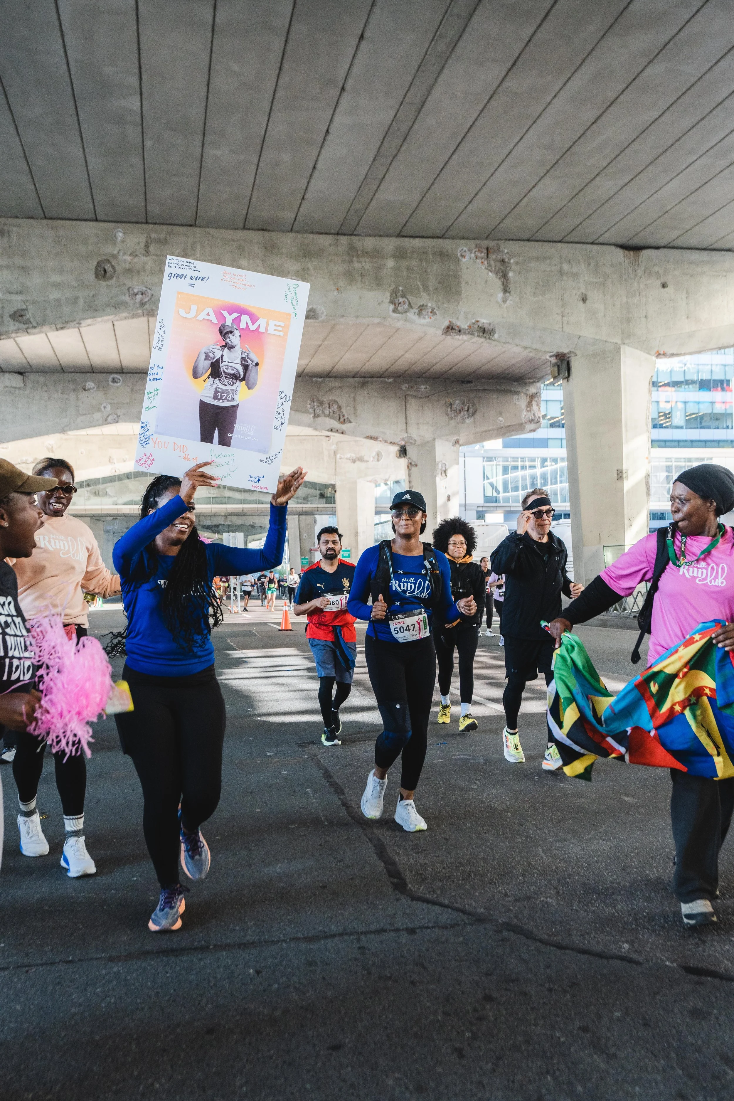 Group of runners participating in a race under a bridge, with one person holding a poster of a woman named Jayme.