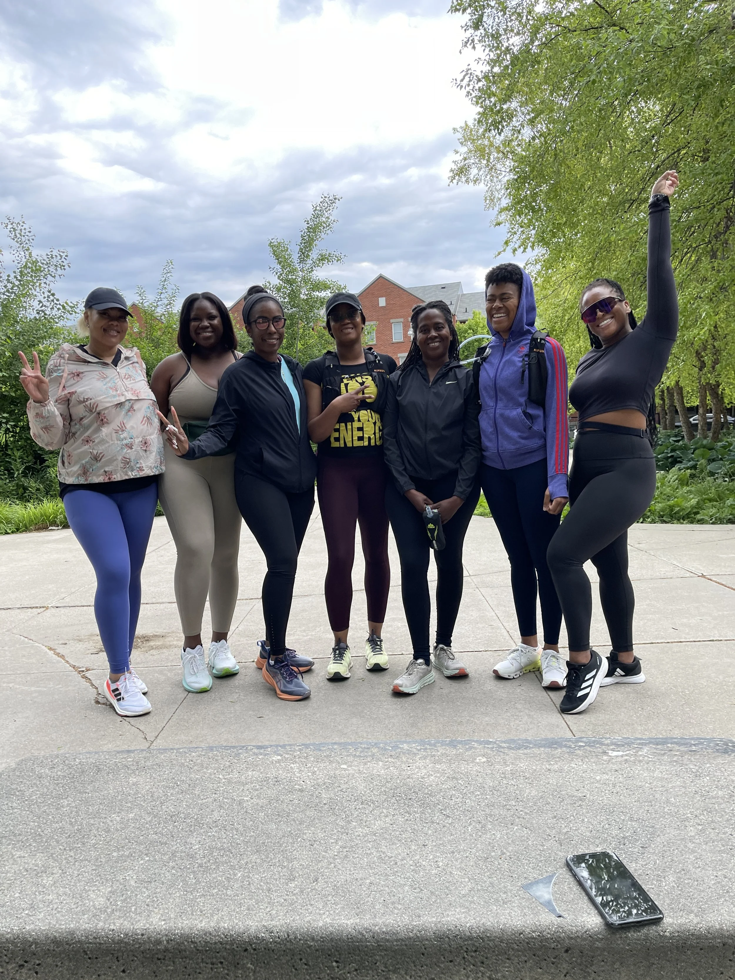 Group of seven women standing outdoors on a paved path, smiling and posing for a photo with trees and houses in the background.