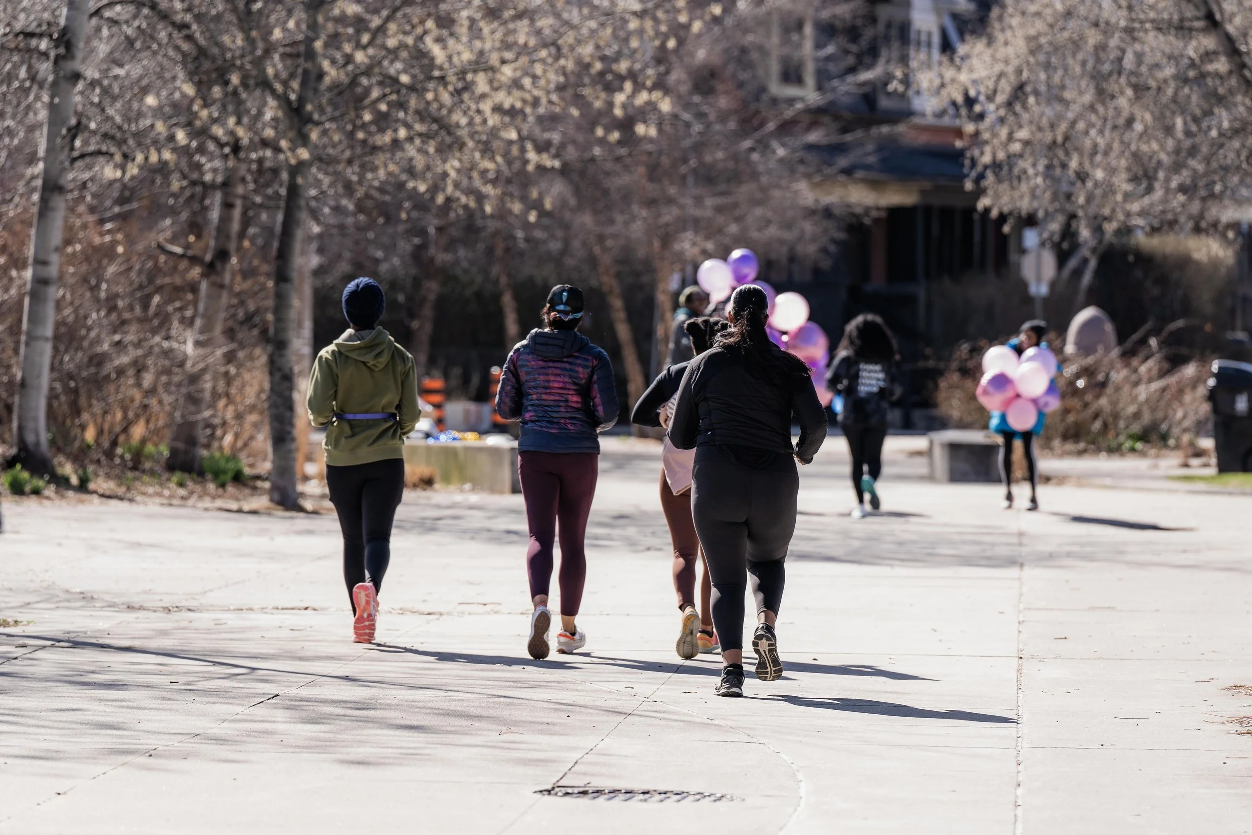 People running outdoors on a sidewalk with trees and balloons in the background.