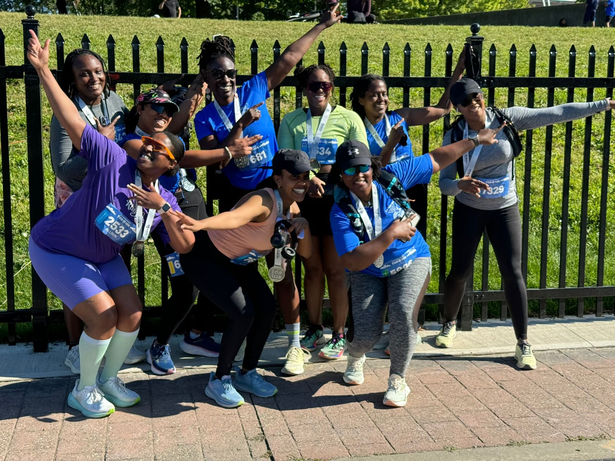 A group of people in athletic clothing and medals around their necks, posing joyful and energetic in front of a black fence, during a running event on a sunny day.