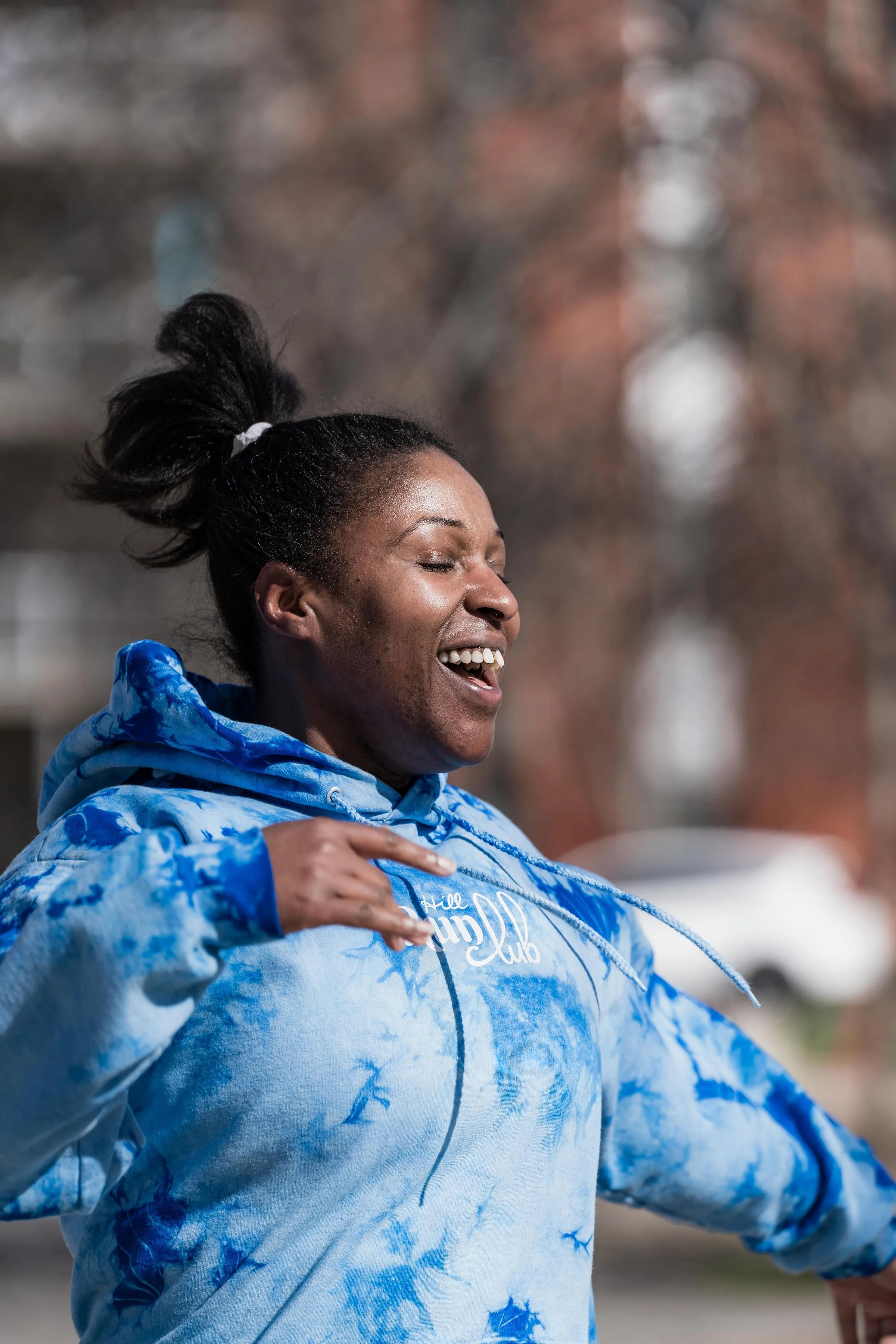 A joyful woman wearing a blue tie-dye hoodie, smiling with eyes closed and hair tied in a high ponytail, outdoors during the daytime.