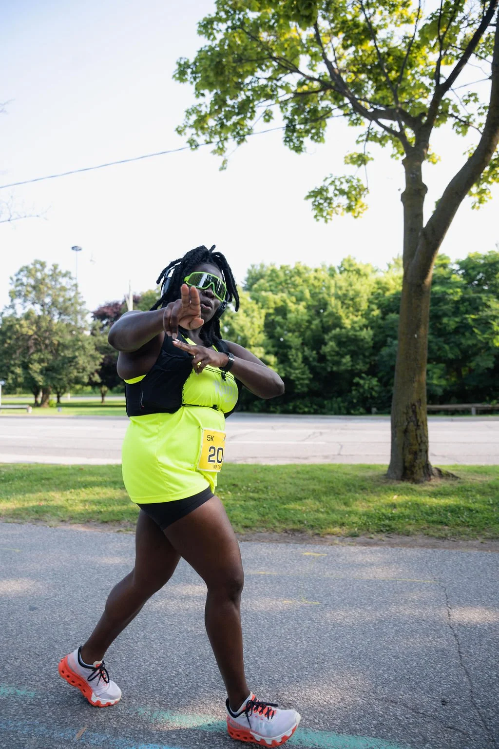 A female runner in neon yellow athletic clothing and sunglasses participating in a 5K race outdoors, with trees and a park in the background.