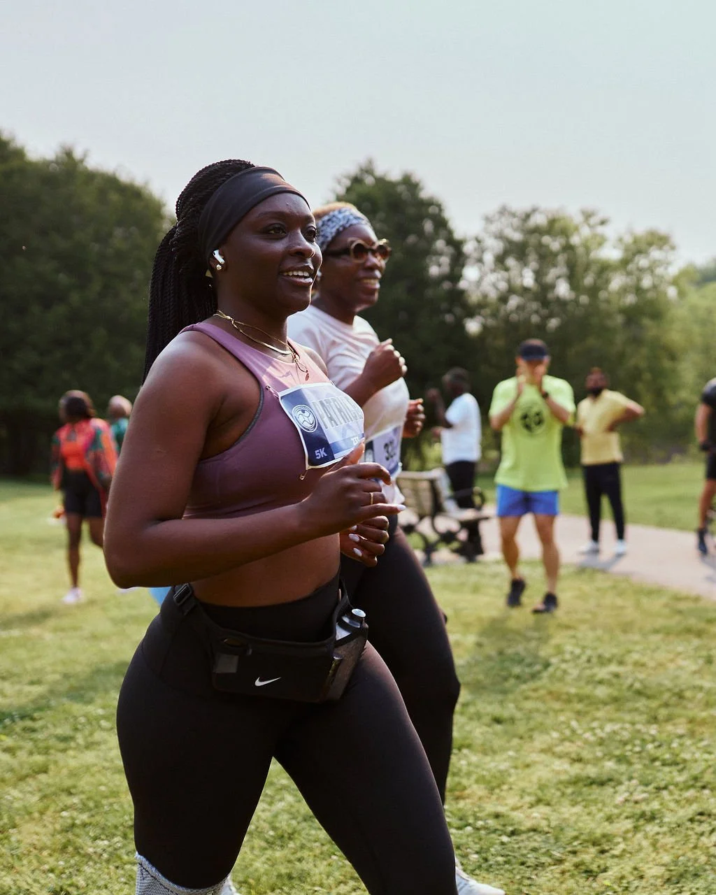 A woman running in a park during a race event, with a smile on her face, wearing a purple sports bra, black leggings, headphones, and a waist pouch, surrounded by other participants and spectators.