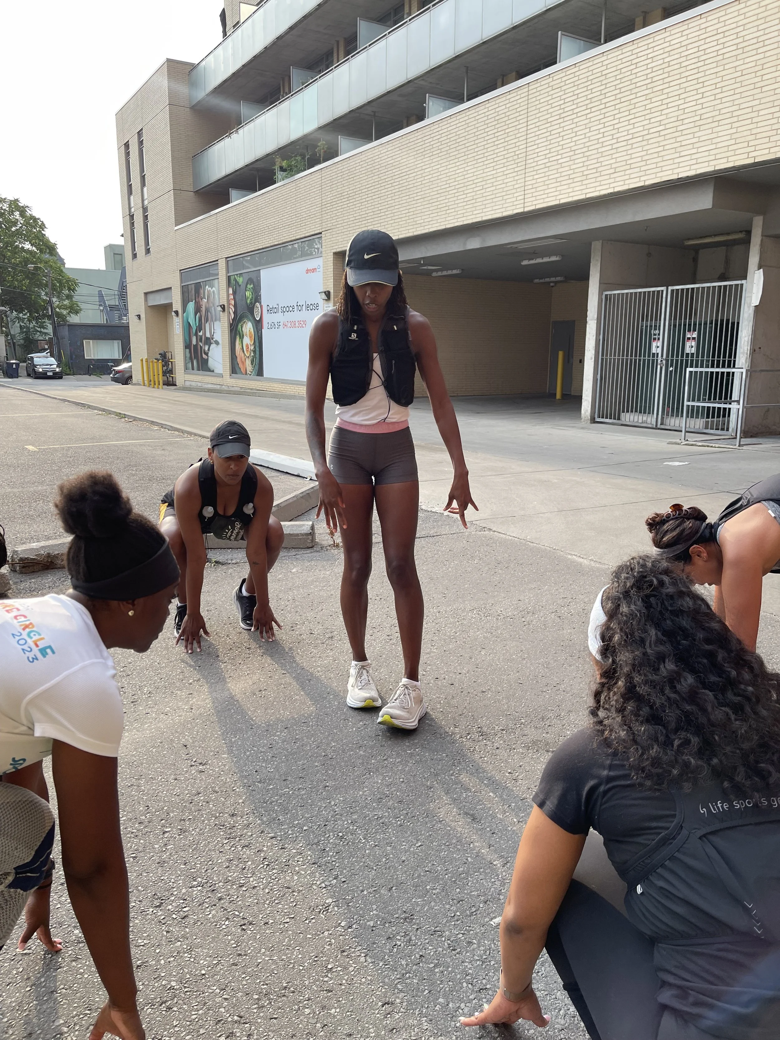 A group of women stretching on an urban street in front of a modern building with glass balconies; one woman is standing, others are crouching or bending down.