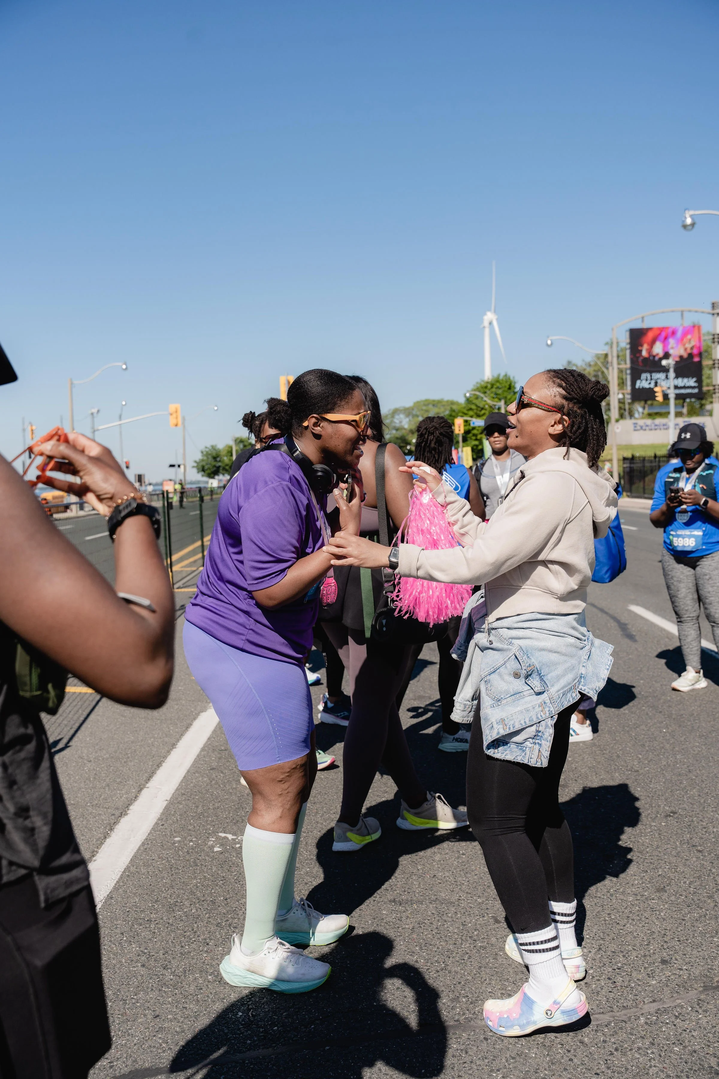Two women are smiling and engaging in friendly conversation on a city street with several other people around, some with bib numbers, indicating a running event or marathon, under clear blue skies.