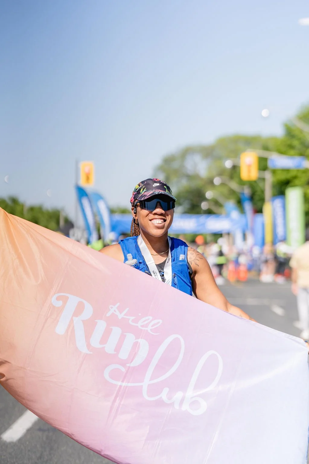 A woman smiling, wearing sunglasses and athletic gear, holding a pink flag with the words 'Hello Rundle' at an outdoor running event on a sunny day.