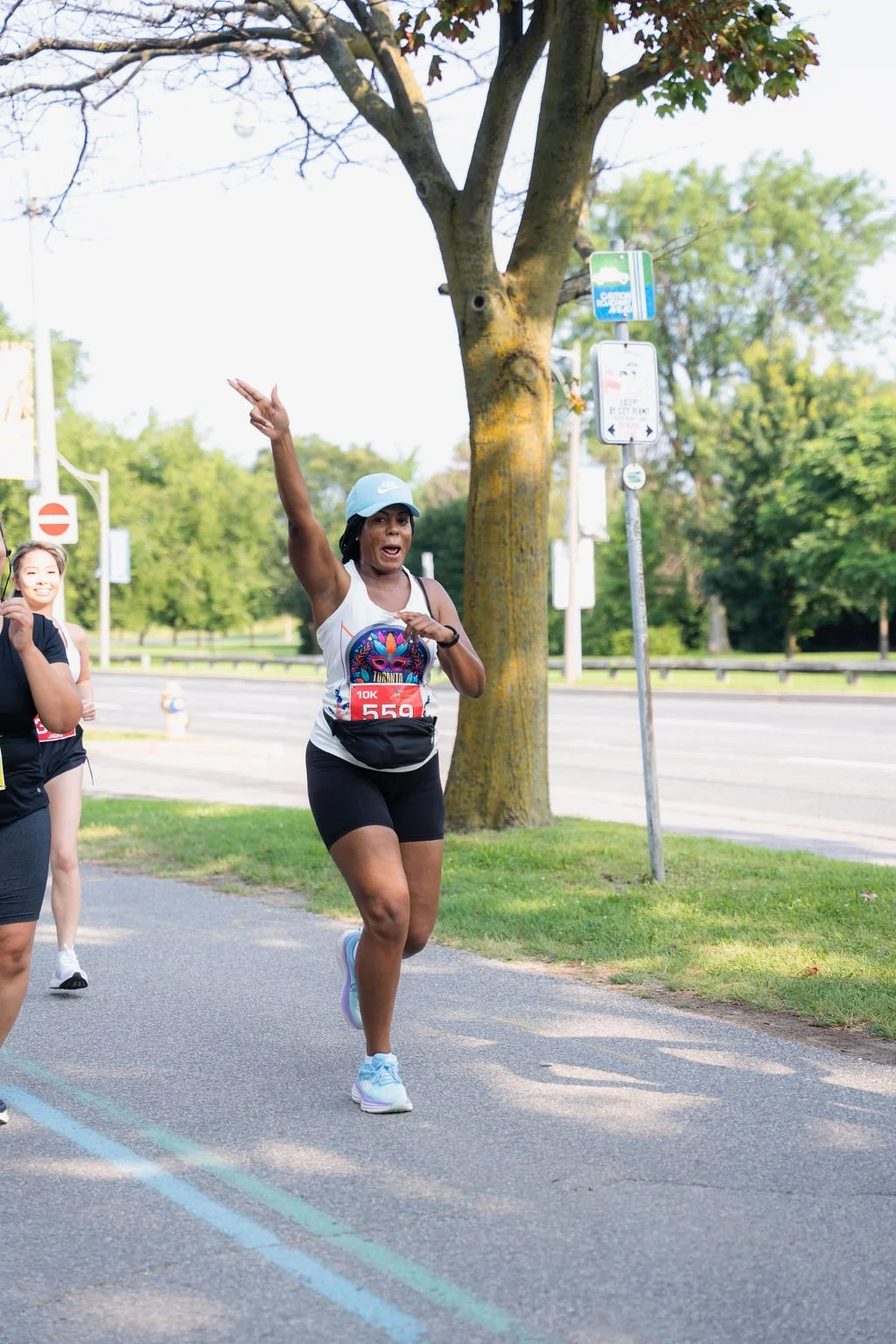 A woman wearing a white tank top, black shorts, and a light blue helmet is running in a race, gesturing with her left hand raised, while smiling and looking ahead. She is running on a paved path with greenery and trees in the background, and other ru