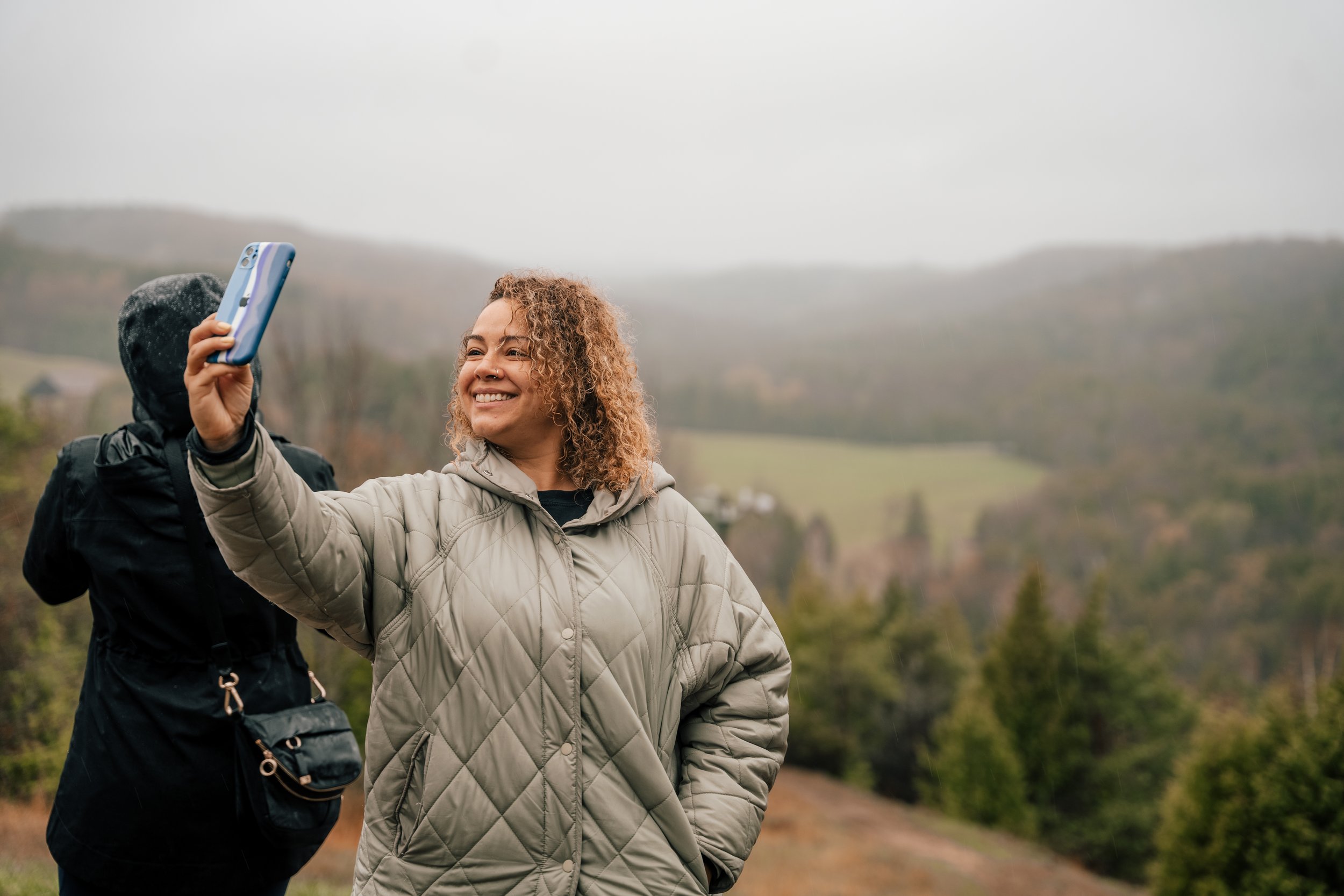 A woman smiling and taking a selfie outdoors on a cloudy day, with rolling hills and trees in the background, and a person in a black jacket standing behind her.