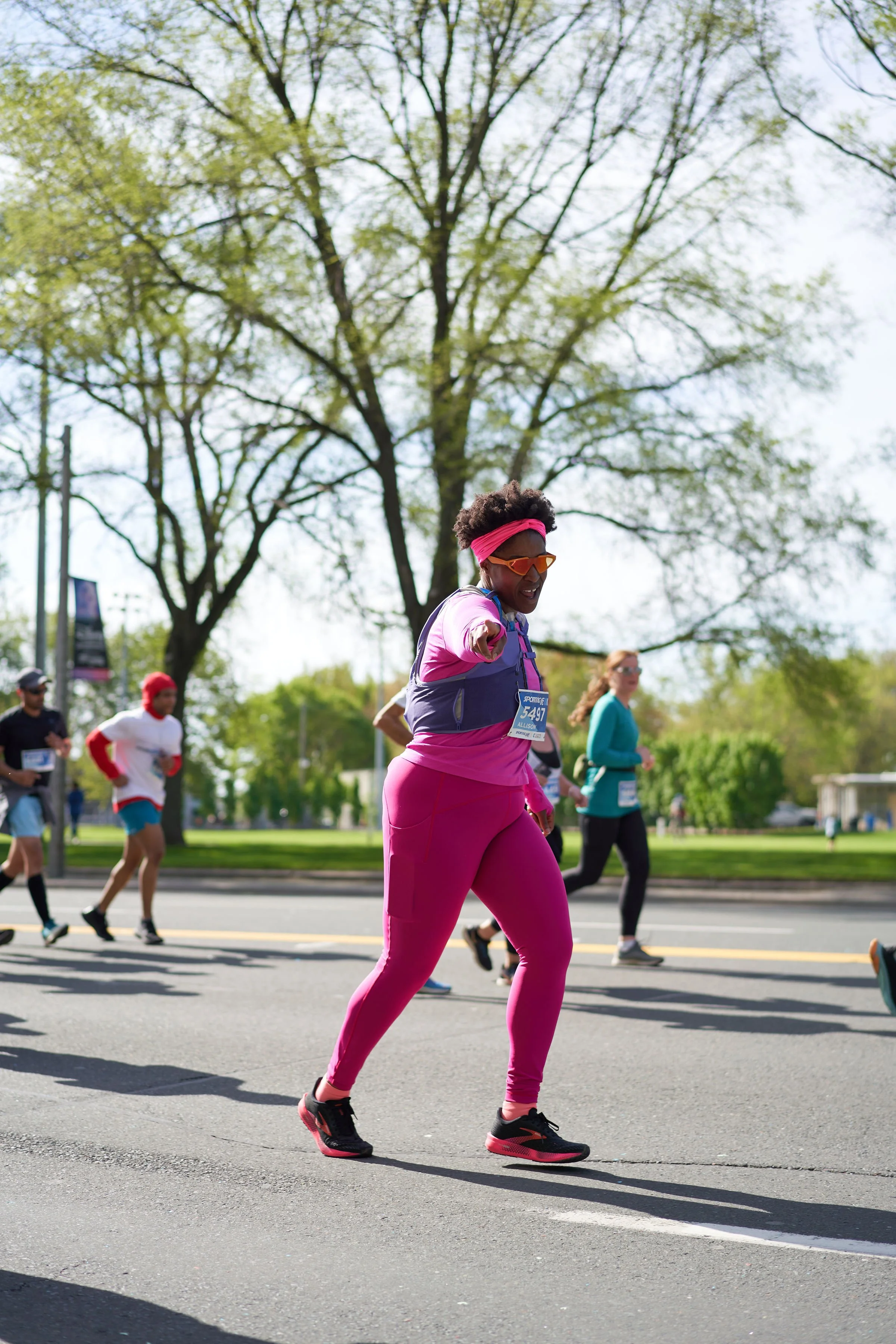 A woman in athletic clothes and sunglasses smiling and pointing toward the camera while running in a marathon. Other runners and leafless trees are visible in the background.