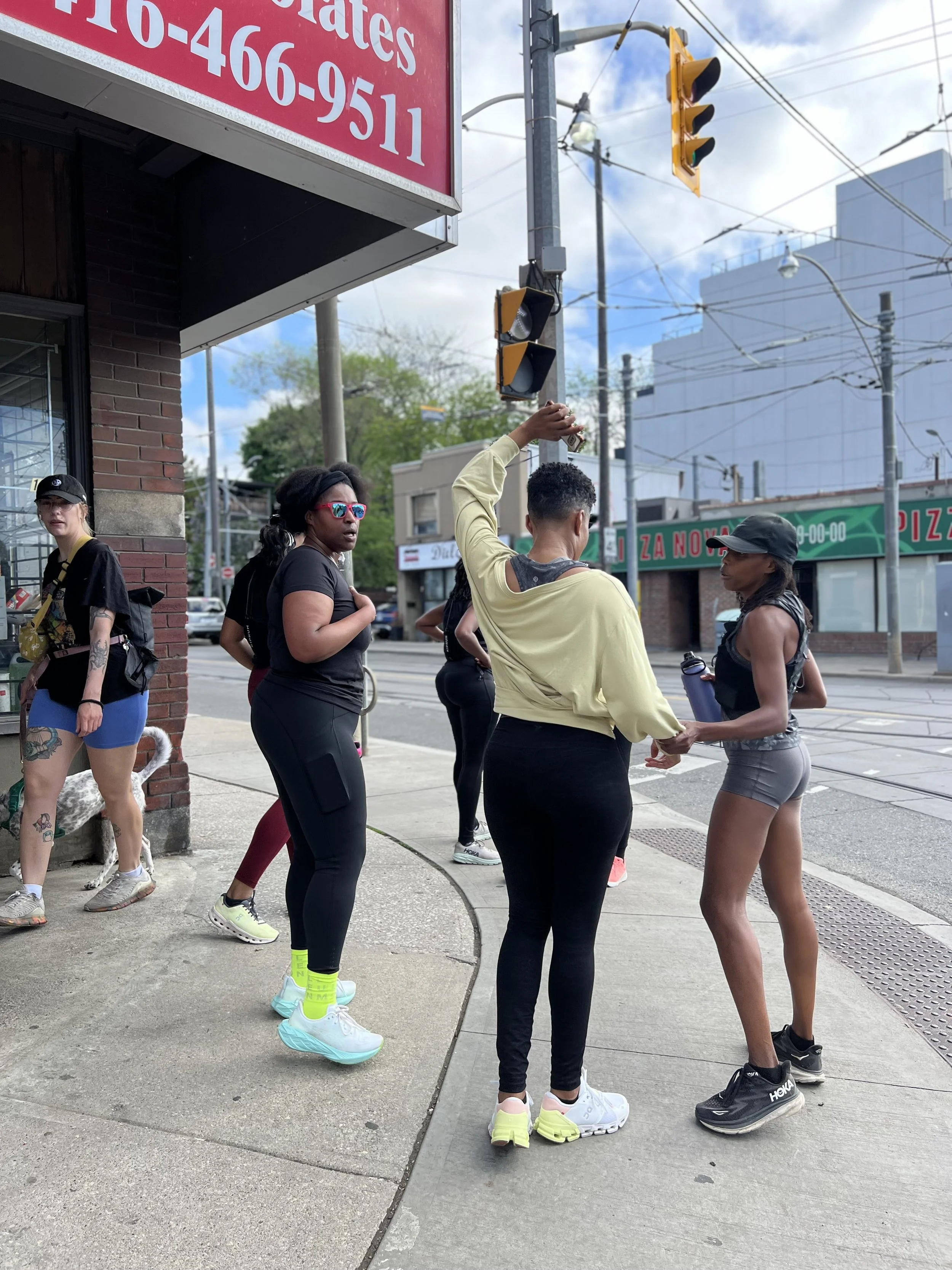 A group of people, mostly women, standing on a city sidewalk at an intersection. Some are talking, and one woman is handing something to another. They are dressed in casual athletic clothing. The background includes traffic lights, power lines, and s