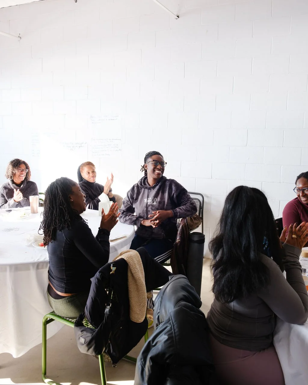 Group of diverse women sitting around a table, laughing and clapping during a social gathering in a room with white brick walls.