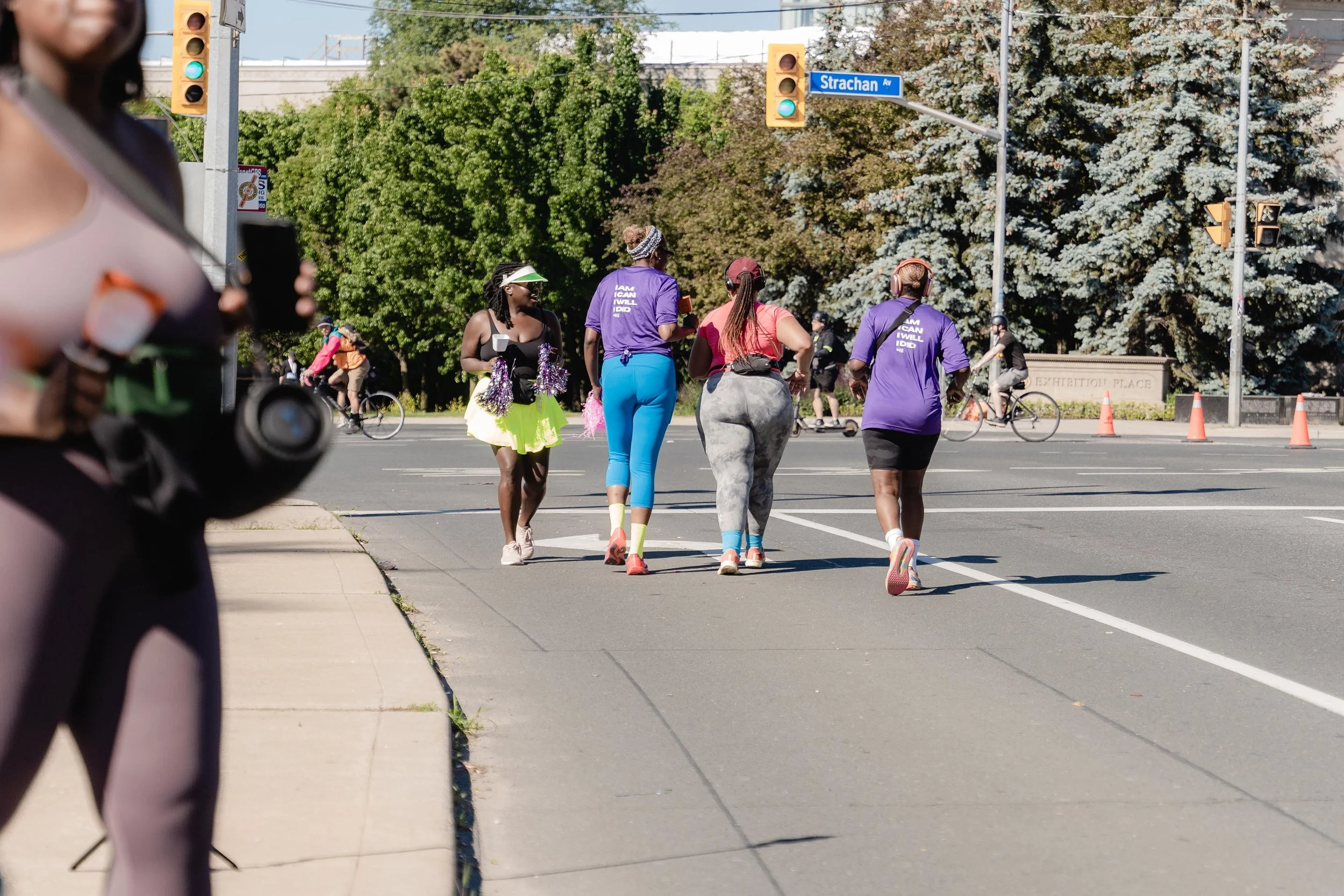 Group of women jogging across a city street, with some cyclists in the background, traffic lights, and trees lining the street.