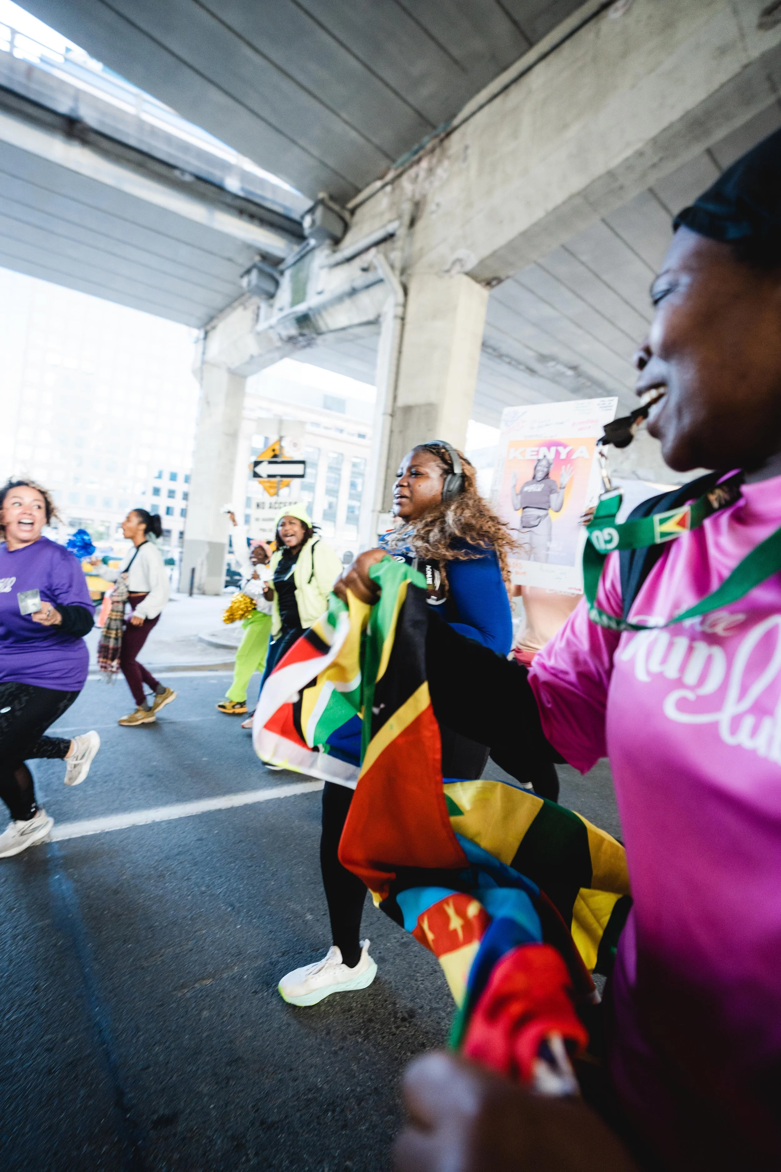 Women participating in a group walk or run event under a bridge, holding flags and posters, some wearing headphones and colorful athletic clothing.