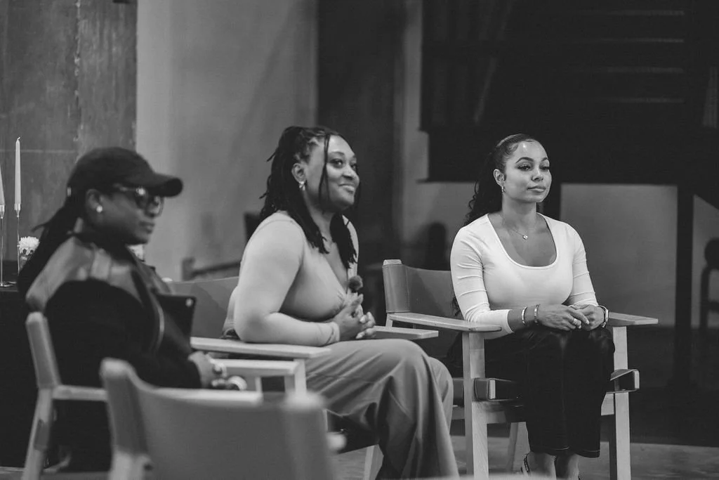 Three women sitting in a panel discussion setting, with one woman smiling and the other two women looking serious, in a black and white photo.
