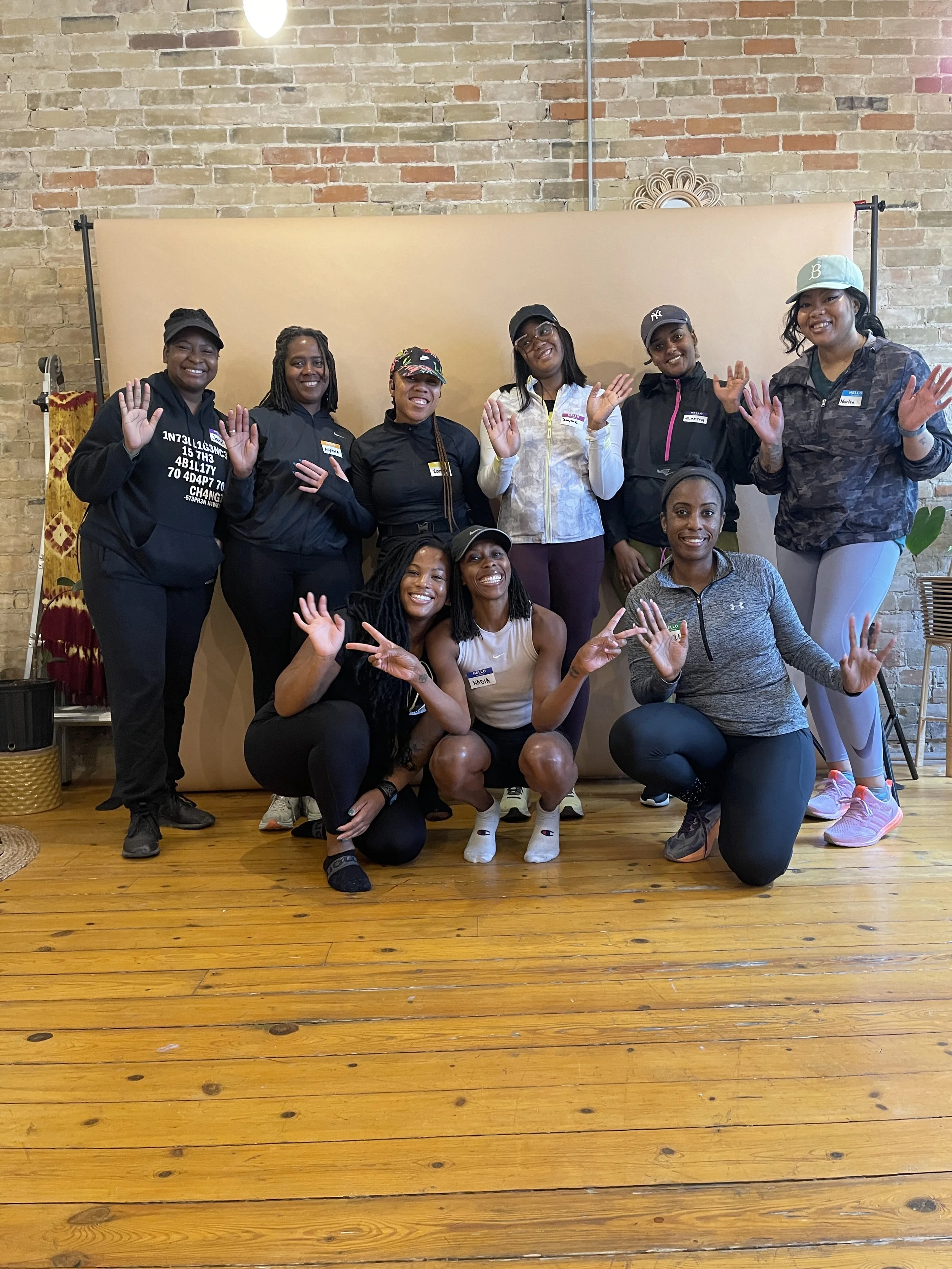 A group of nine women posing together indoors with a beige backdrop, smiling and waving at the camera.