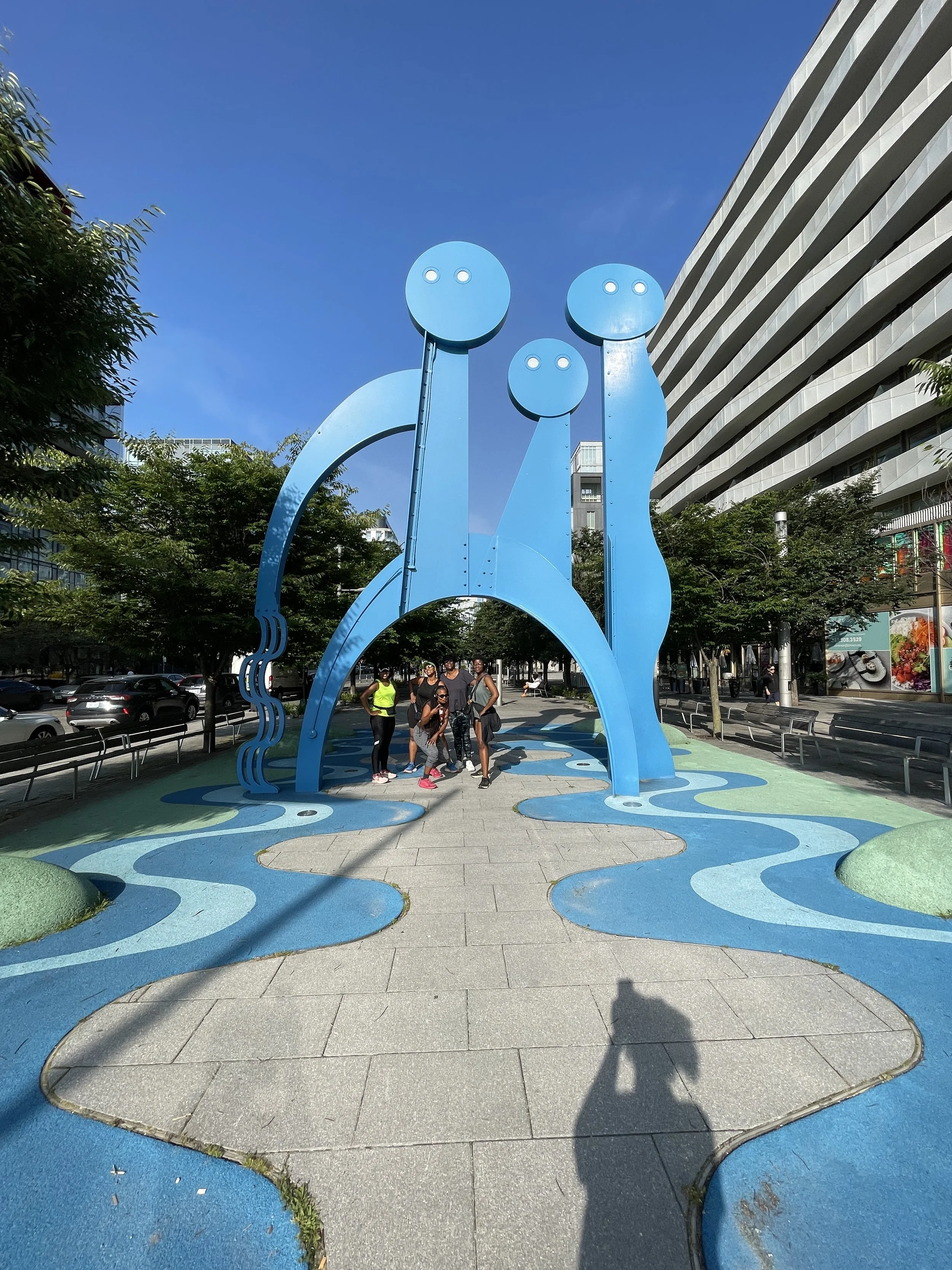 Blue sculpture of three abstract human figures with circular heads and elongated bodies, situated in an urban park, with people walking underneath. The ground is decorated with blue and green wavy patterns.