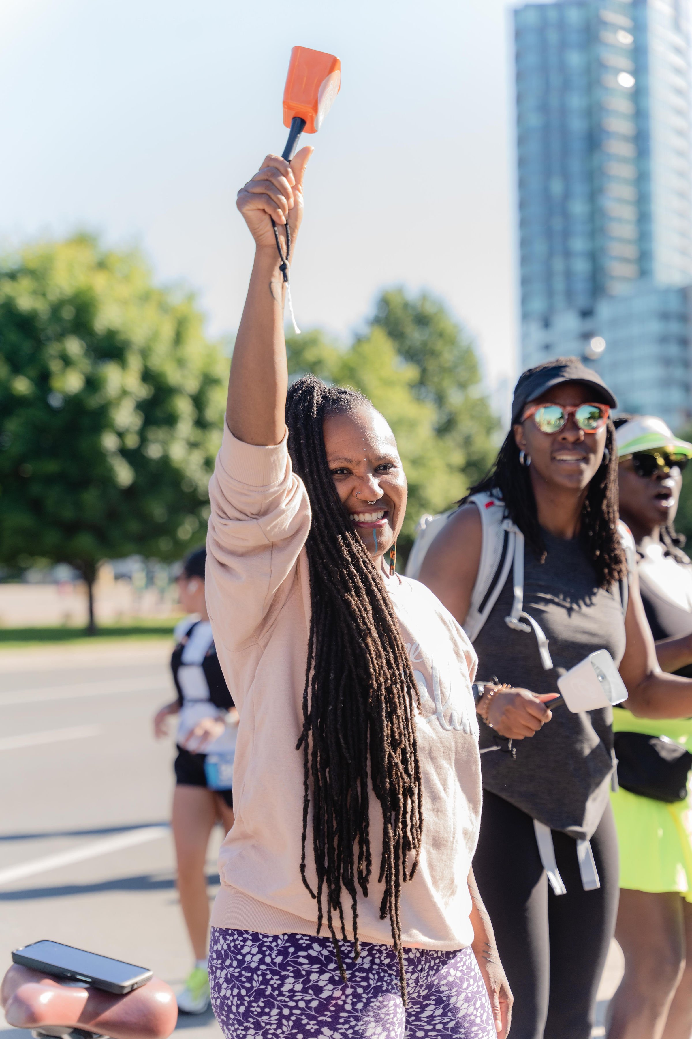 A woman with long dreadlocks holding a whistle up in the air during an outdoor event, with other women in athletic attire and sunglasses in the background.