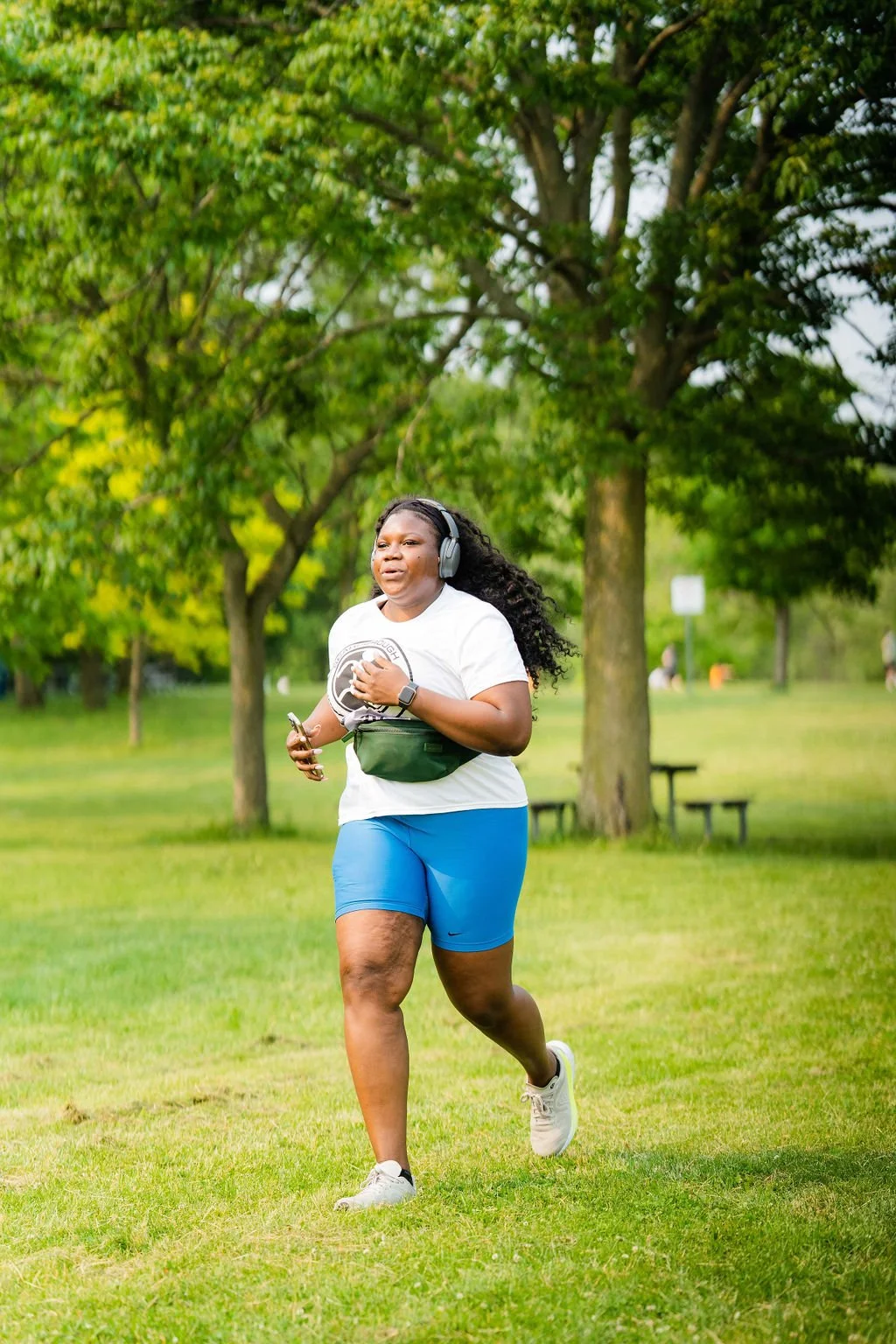 A woman jogs in a park with green trees in the background, wearing a white t-shirt, blue shorts, and white sneakers, with headphones, a watch, and holding a smartphone.