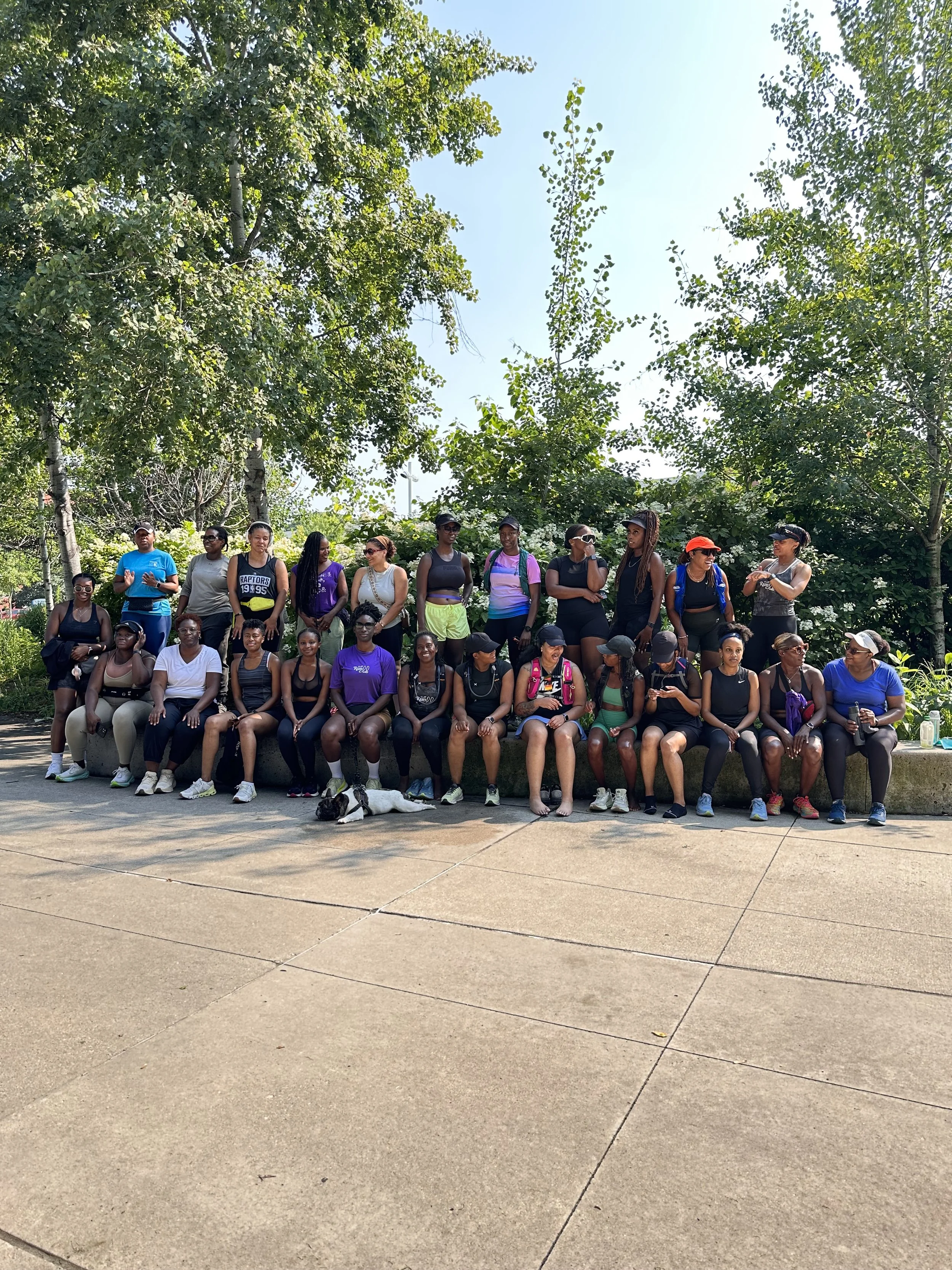 Group of women sitting and standing outdoors on a concrete area, with trees and clear sky in the background.