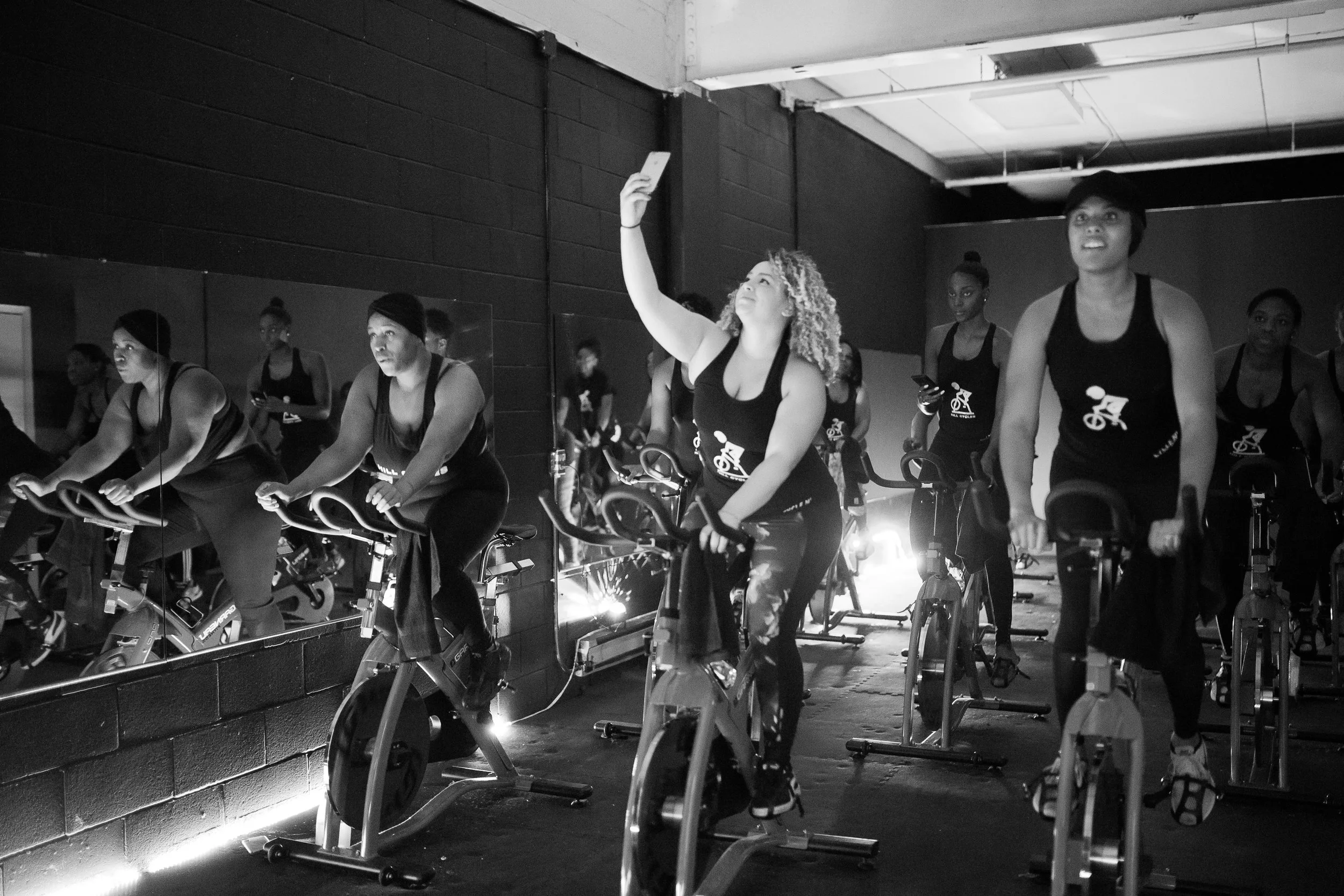 A group of women participating in a spin class in a fitness studio, with some using smartphones, one woman taking a selfie, and a mirrored wall reflecting their movements.