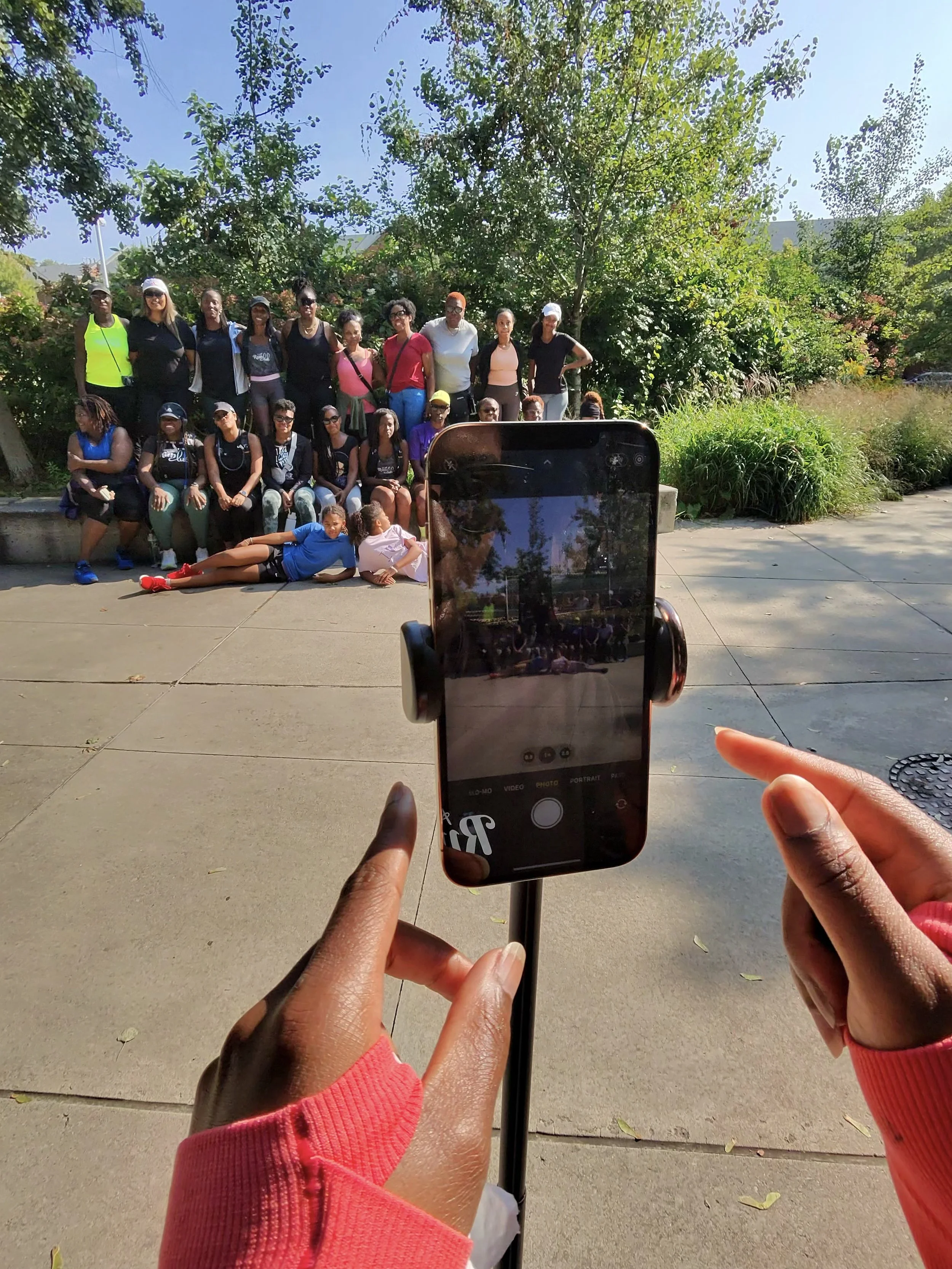 A group of people posing outdoors in front of trees, with someone taking a photo of them with a smartphone.