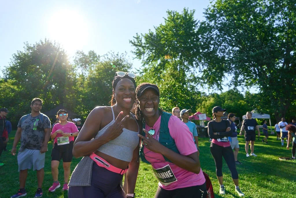 Two women smiling and posing after a race, standing in a park with other runners and trees in the background, with the sun shining.