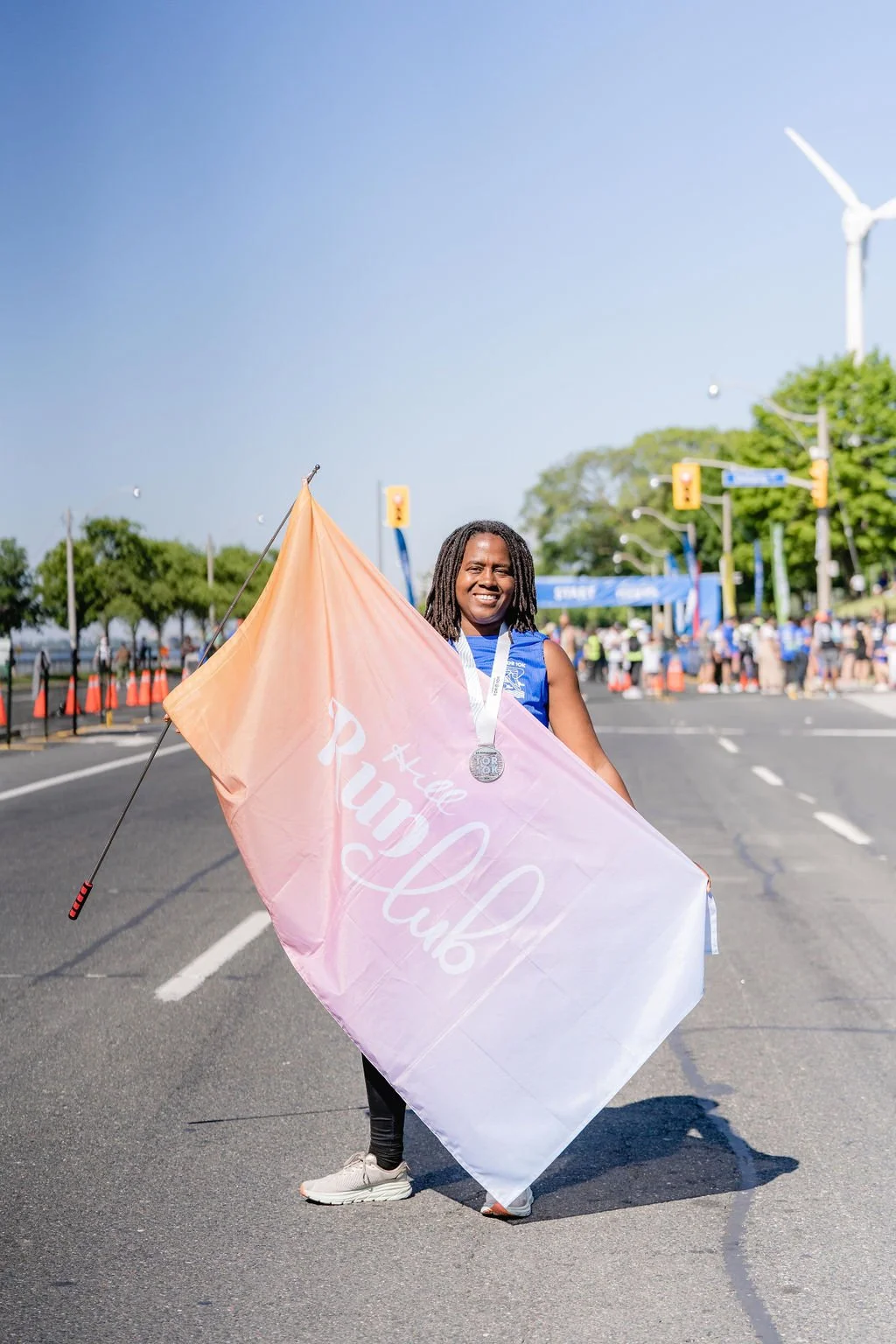 A smiling woman with a medal around her neck holding a pink flag that reads 'Ace the Pud Cup' on a city street during a race or marathon, with other participants and a wind turbine in the background.