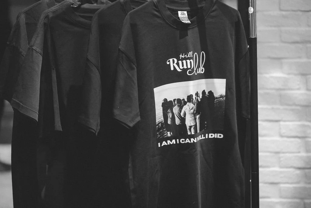 Black T-shirts hanging on a rack, with one shirt displaying a graphic and text that reads "Hill Rum club" and "I am confident I did". The shirt shows a photo of a group of people outdoors near a body of water.