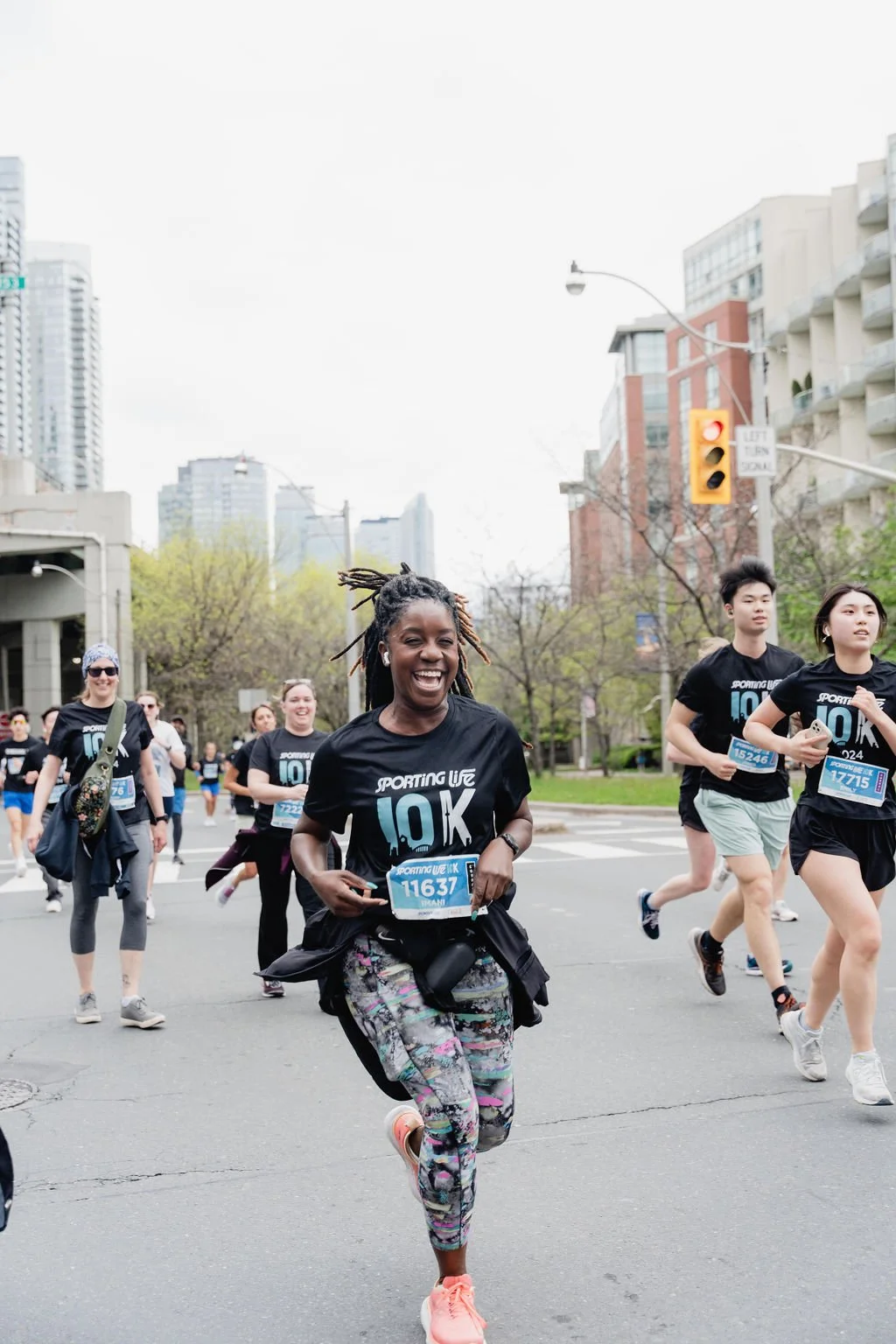 A group of people running in a 10K race on a city street, with a smiling woman in the foreground wearing a black t-shirt with 'SPORTING LIFE 10K' and colorful leggings.