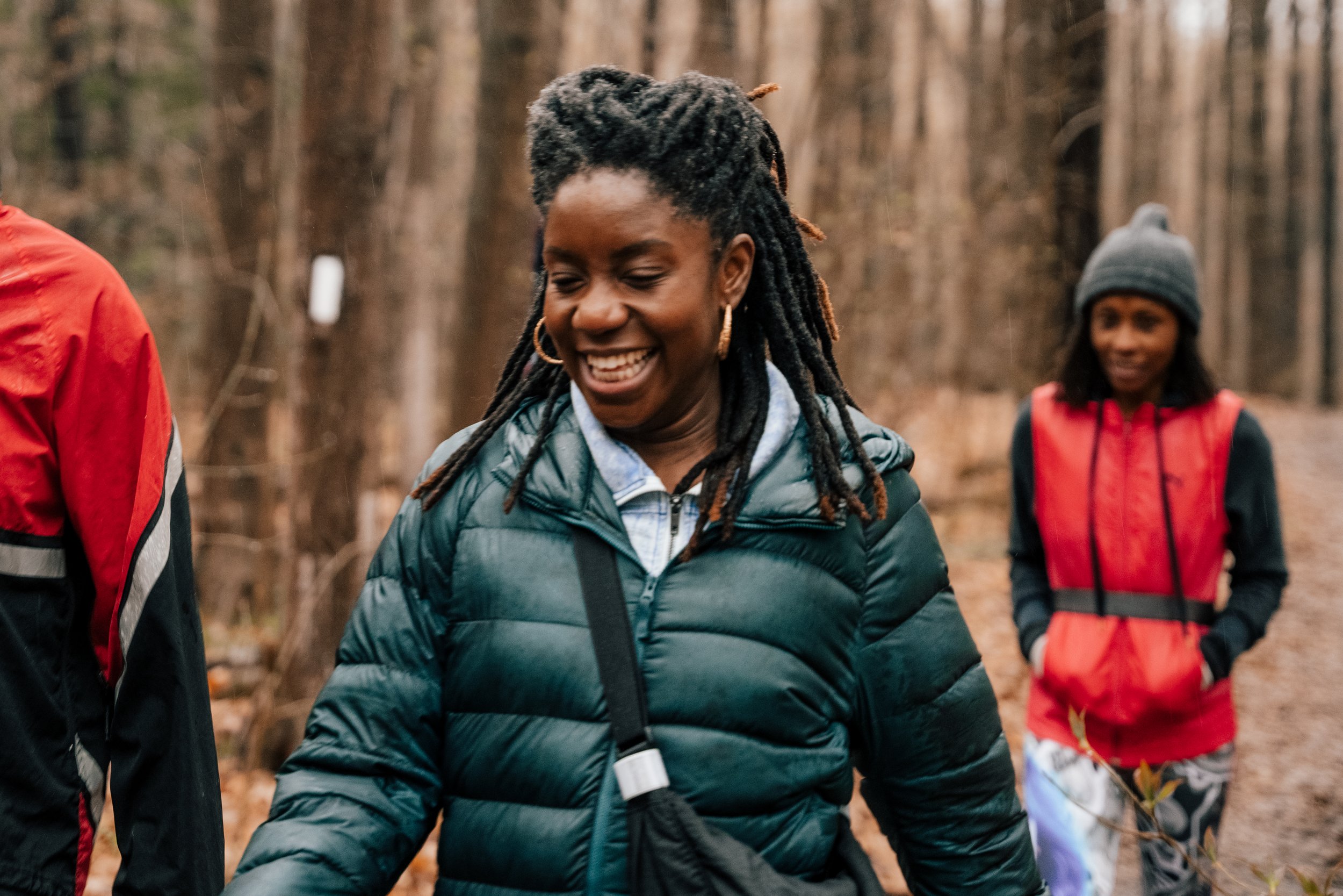 Group of women walking in a wooded outdoor area, smiling, wearing jackets and casual clothing.